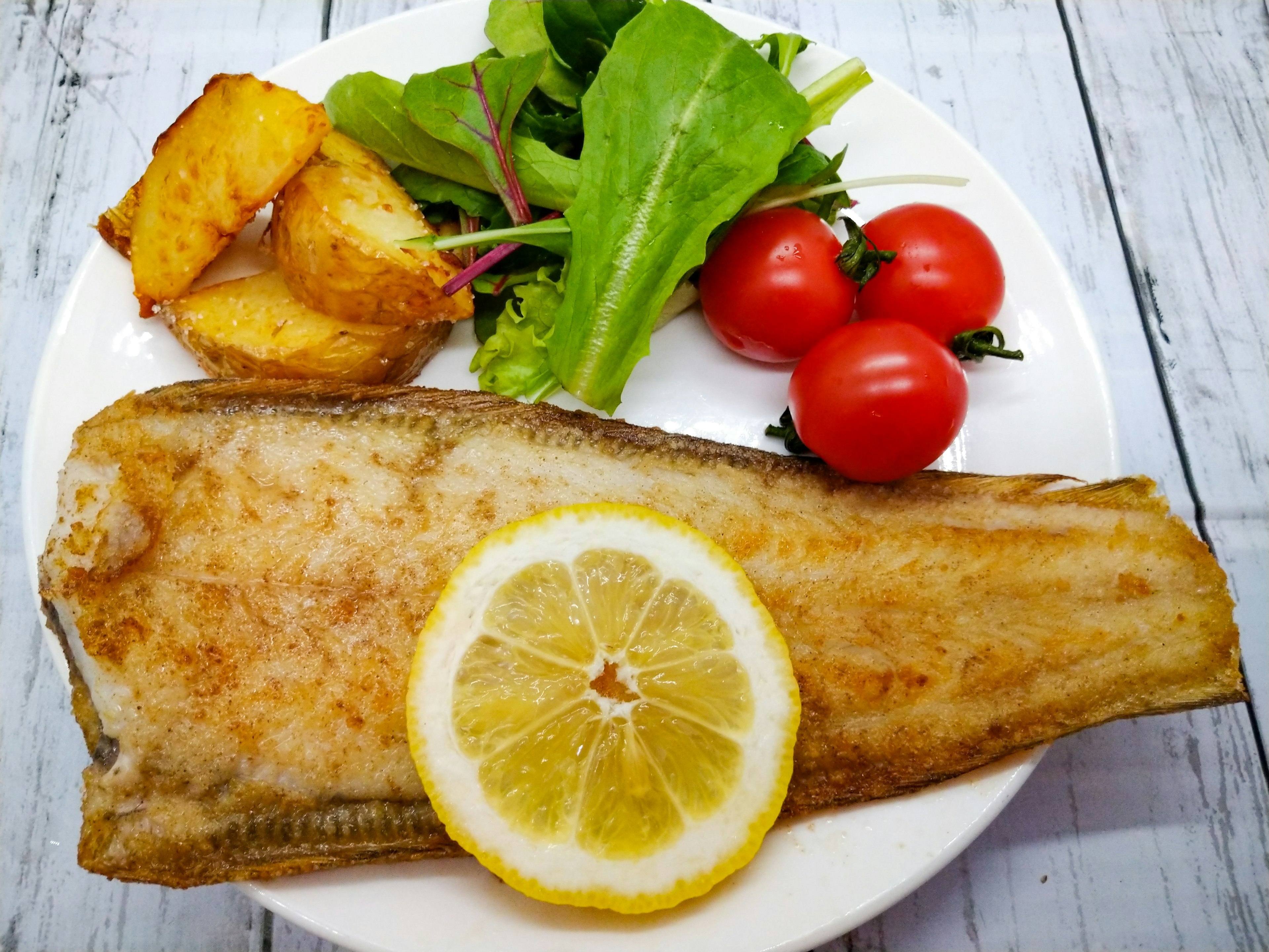 Teller mit Fischfilet garniert mit Zitronenscheibe Salat Kirschtomaten und gerösteten Kartoffeln