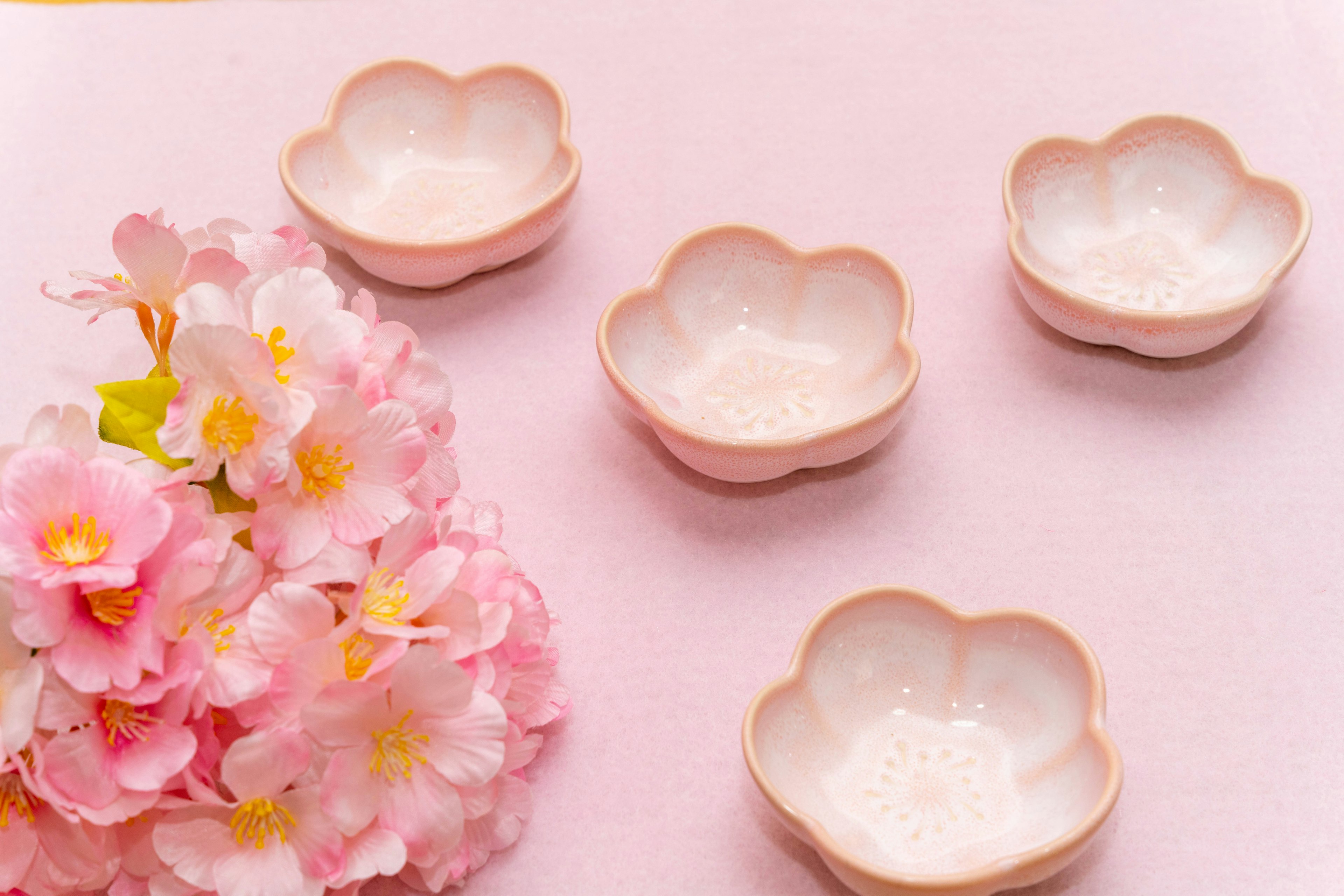 Cuatro pequeños cuencos en forma de pétalo dispuestos sobre un fondo rosa con flores de cerezo rosas