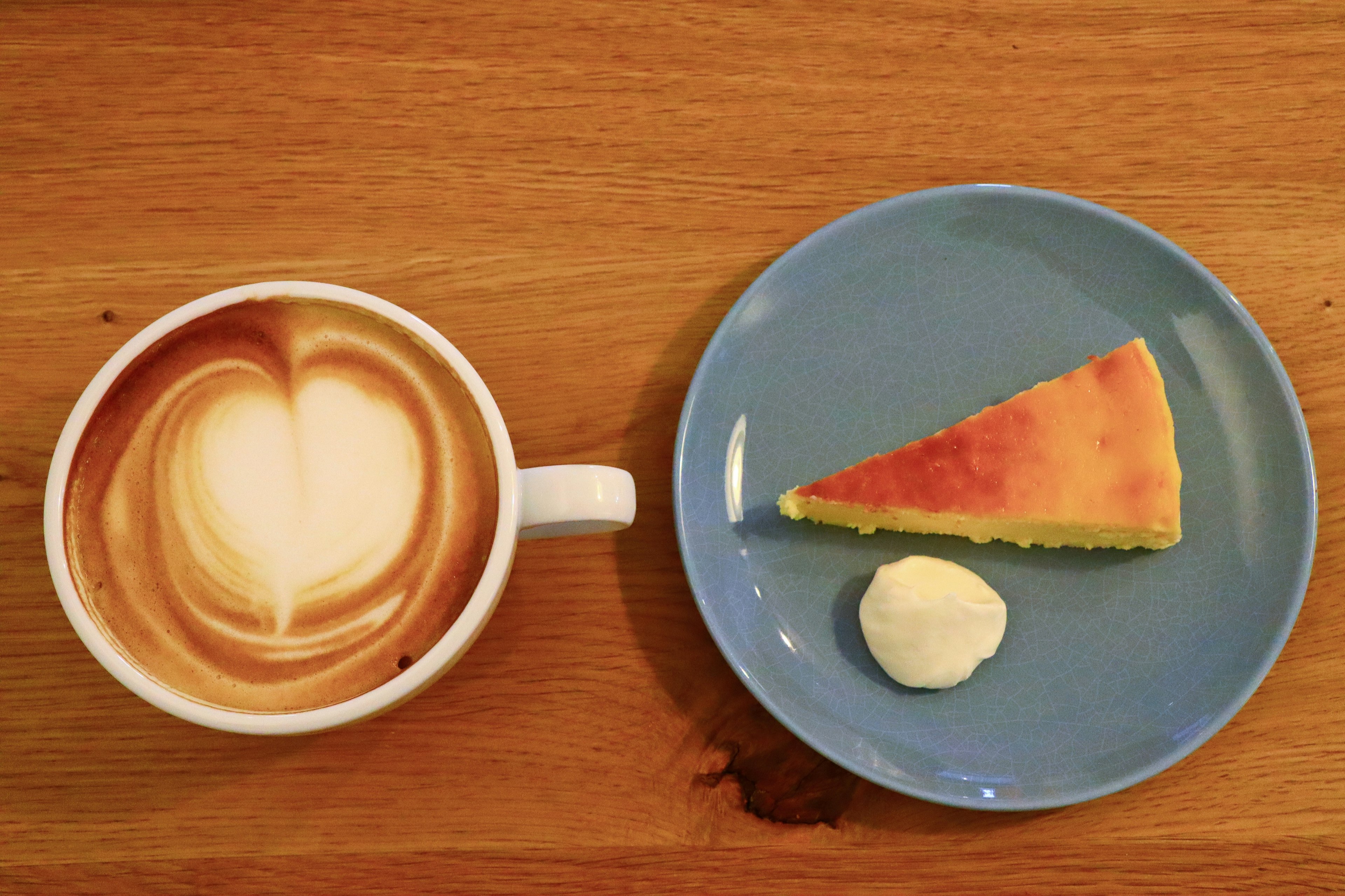 Sebuah latte dengan desain hati dan sepotong kue di piring biru di atas meja kayu