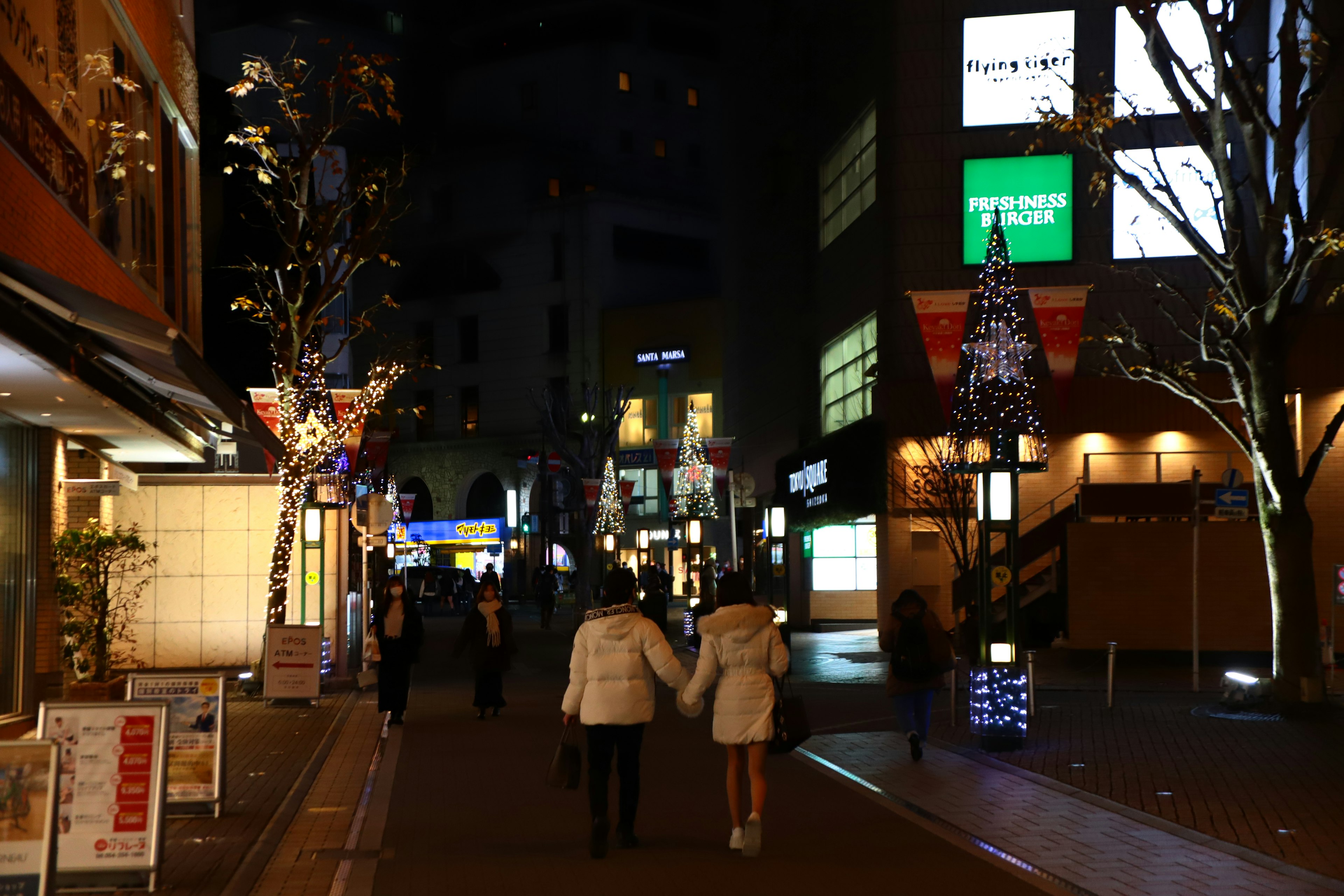 คู่รักเดินในถนนยามค่ำคืนมีต้นไม้และร้านค้าที่สว่างไสว