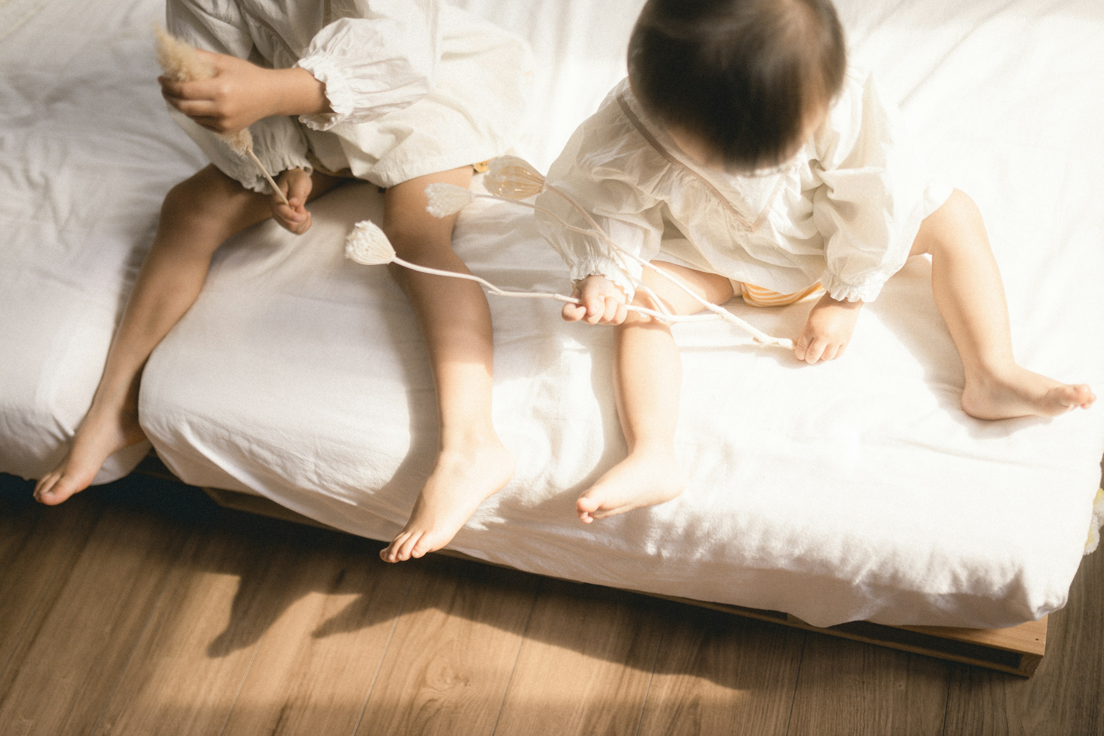 Zwei Kinder spielen auf einem Bett mit Sonnenlicht, das hereinfällt Weiche weiße Bettwäsche und sichtbarer Holzboden