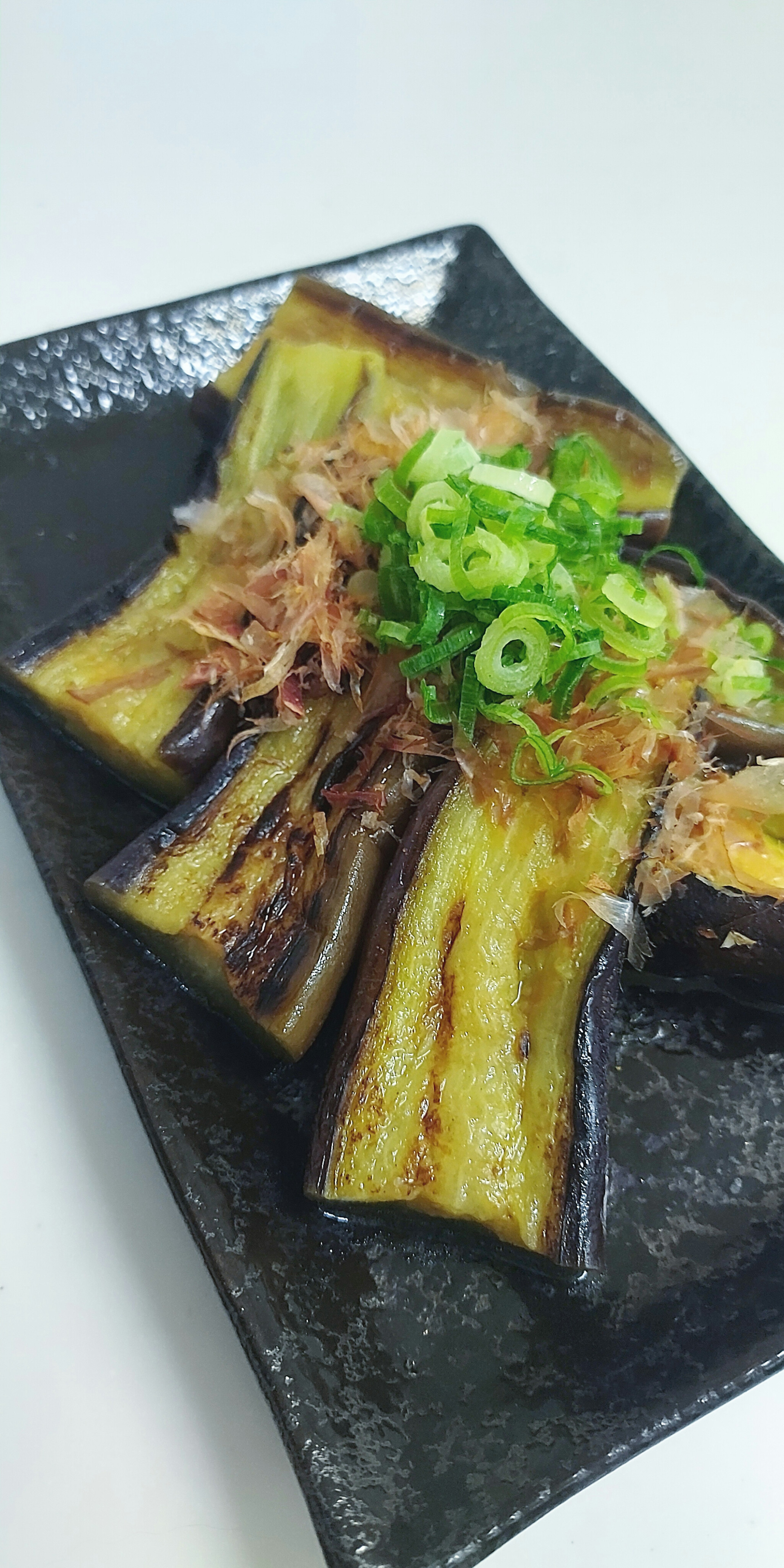 Plato de berenjena a la parrilla servido en un plato negro adornado con cebollas verdes y escamas de bonito