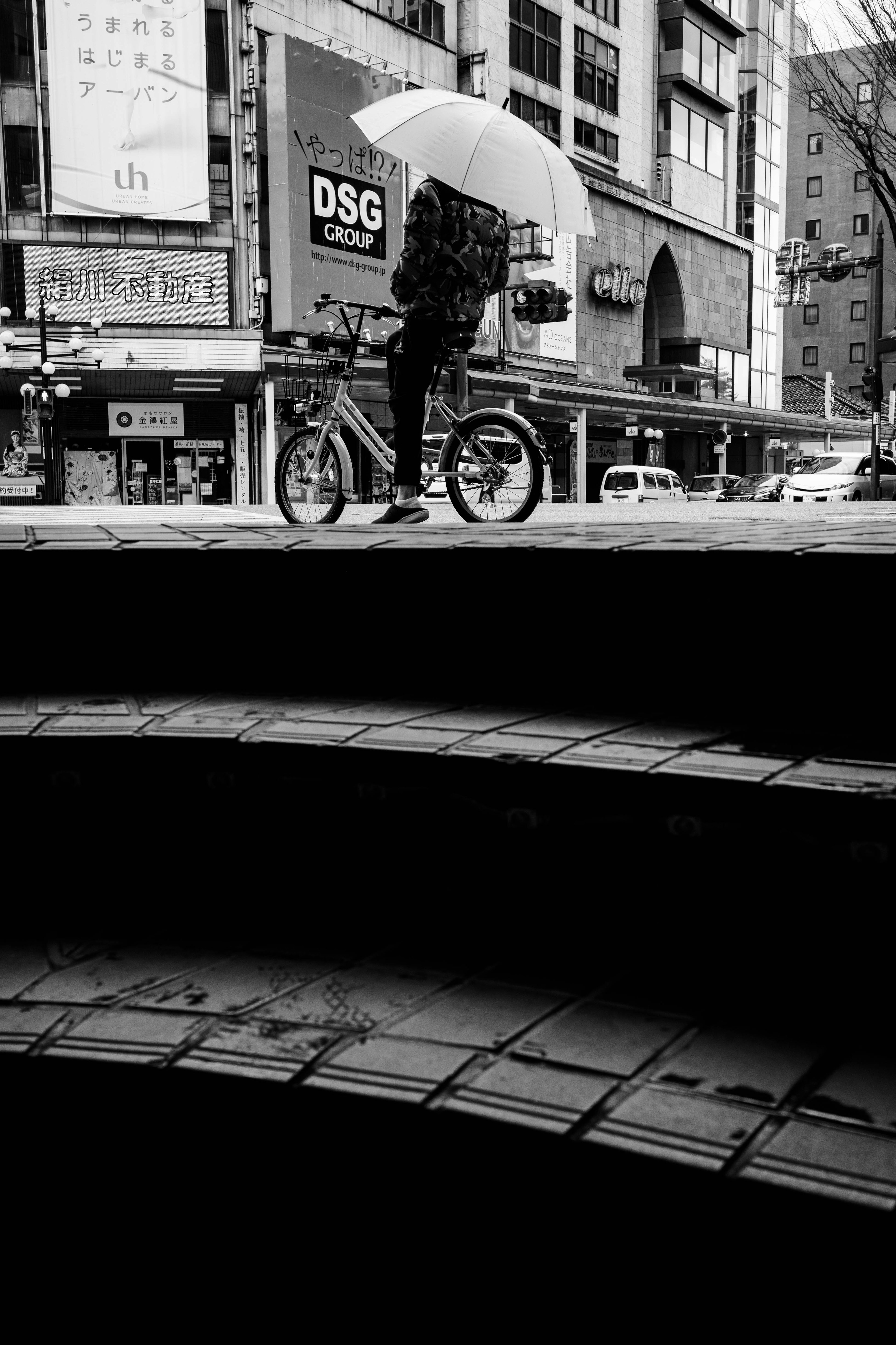 Black and white image of a person on a bicycle holding an umbrella with urban scenery and stairs