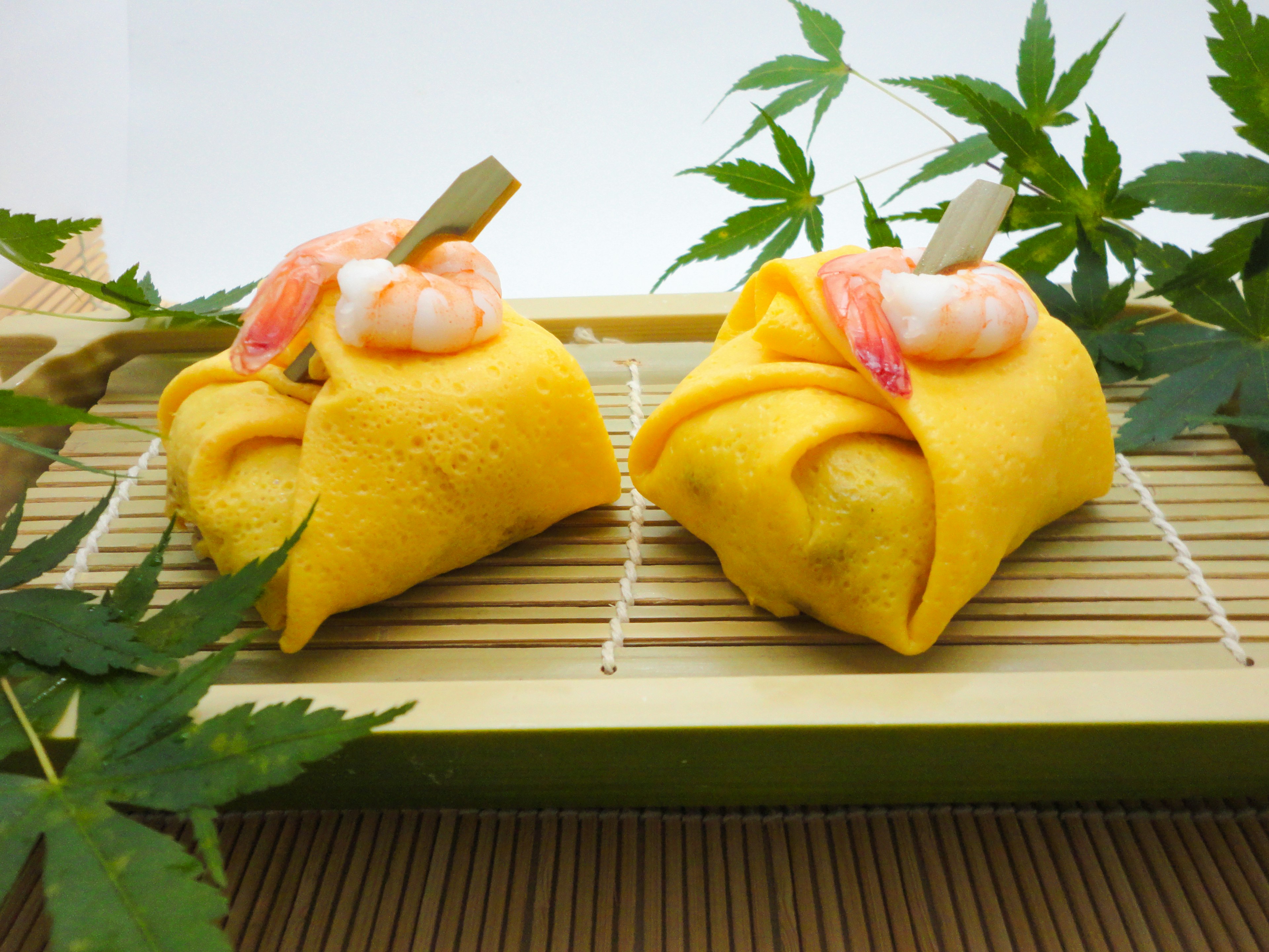 Dos dumplings amarillos con camarones encima dispuestos sobre una estera de bambú rodeados de hojas verdes