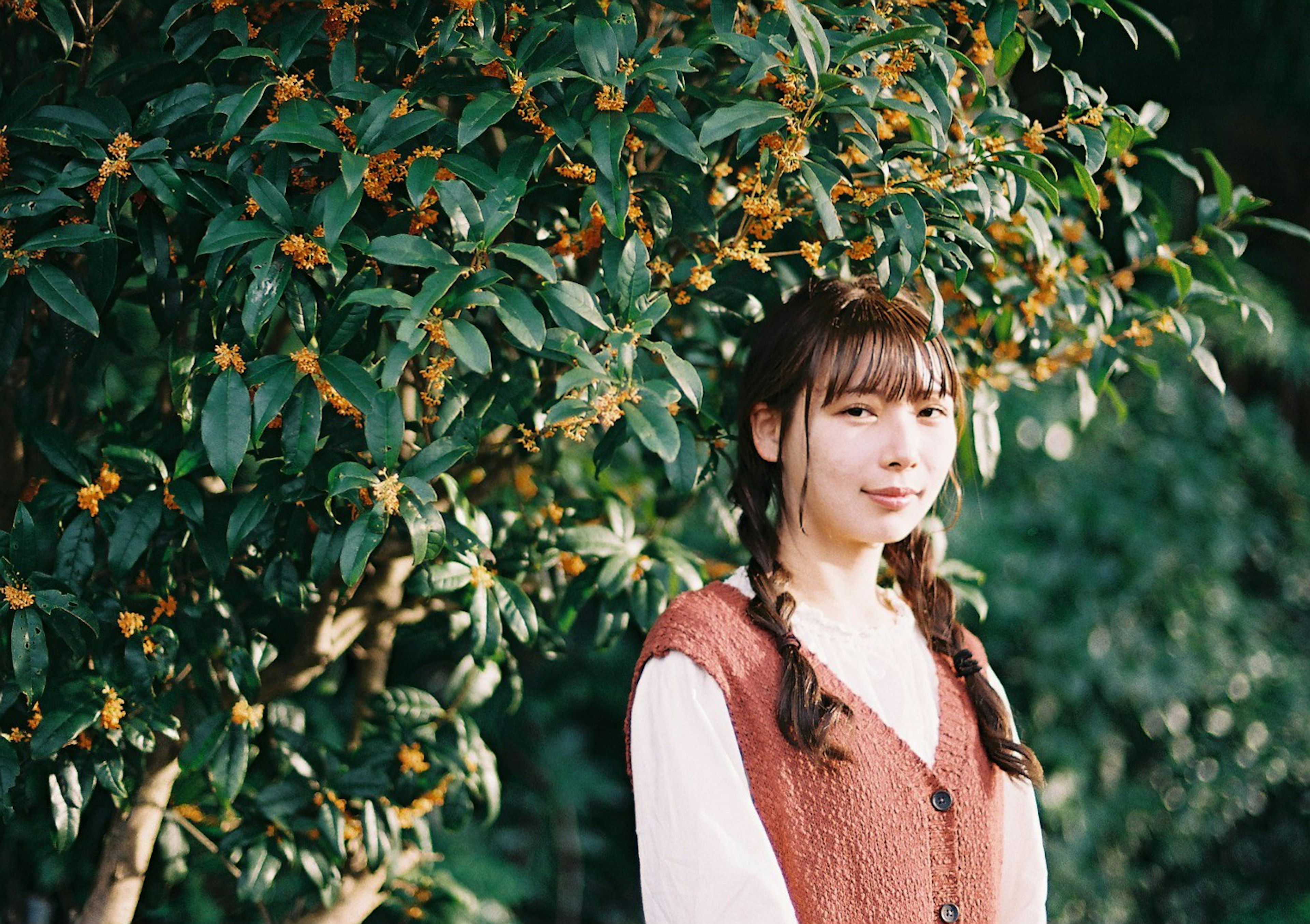 A woman standing in front of a green tree wearing a brown vest with braided hair