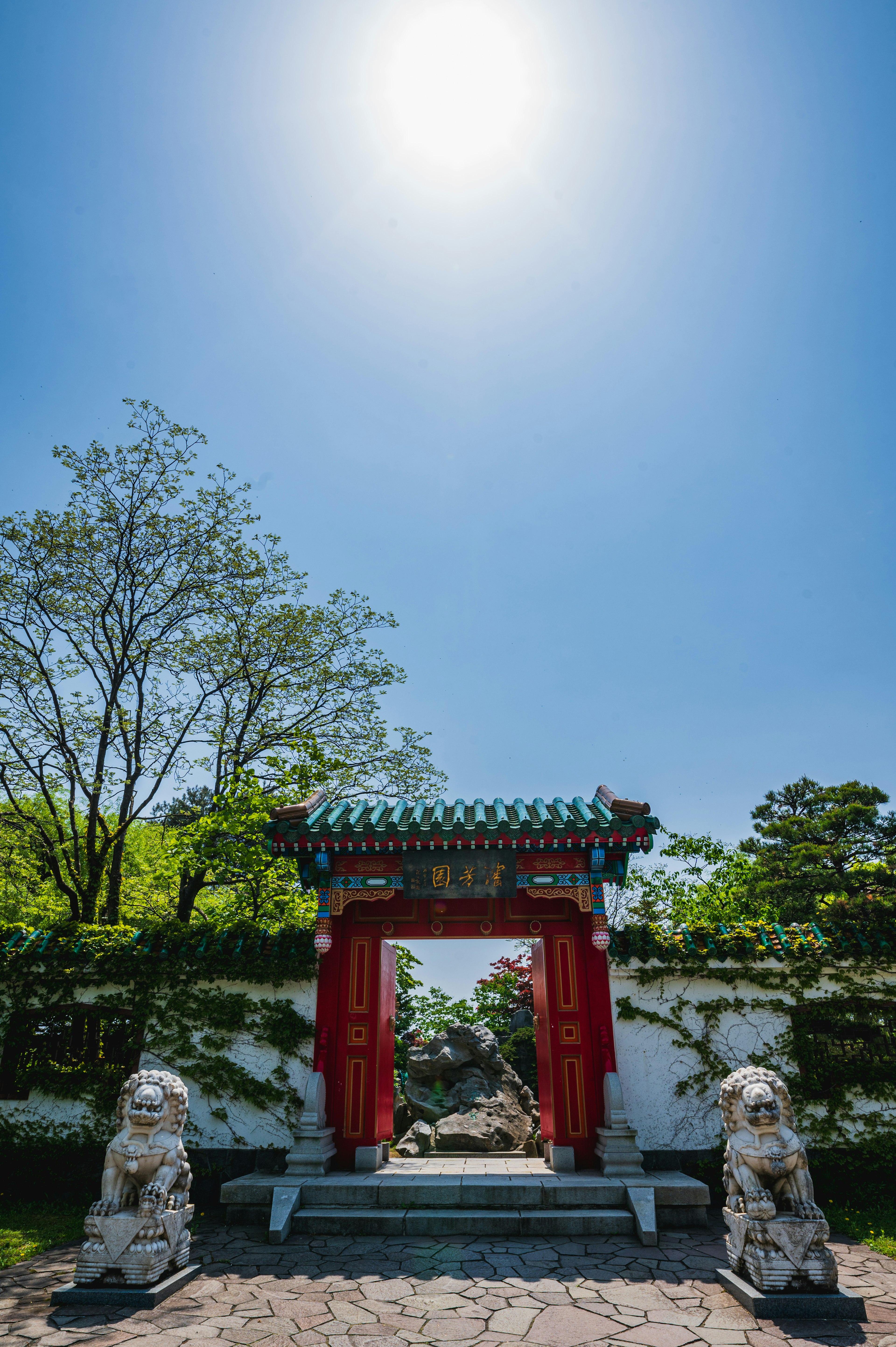 Entrada de jardín chino con una puerta roja y esculturas de leones de piedra bajo un cielo azul claro