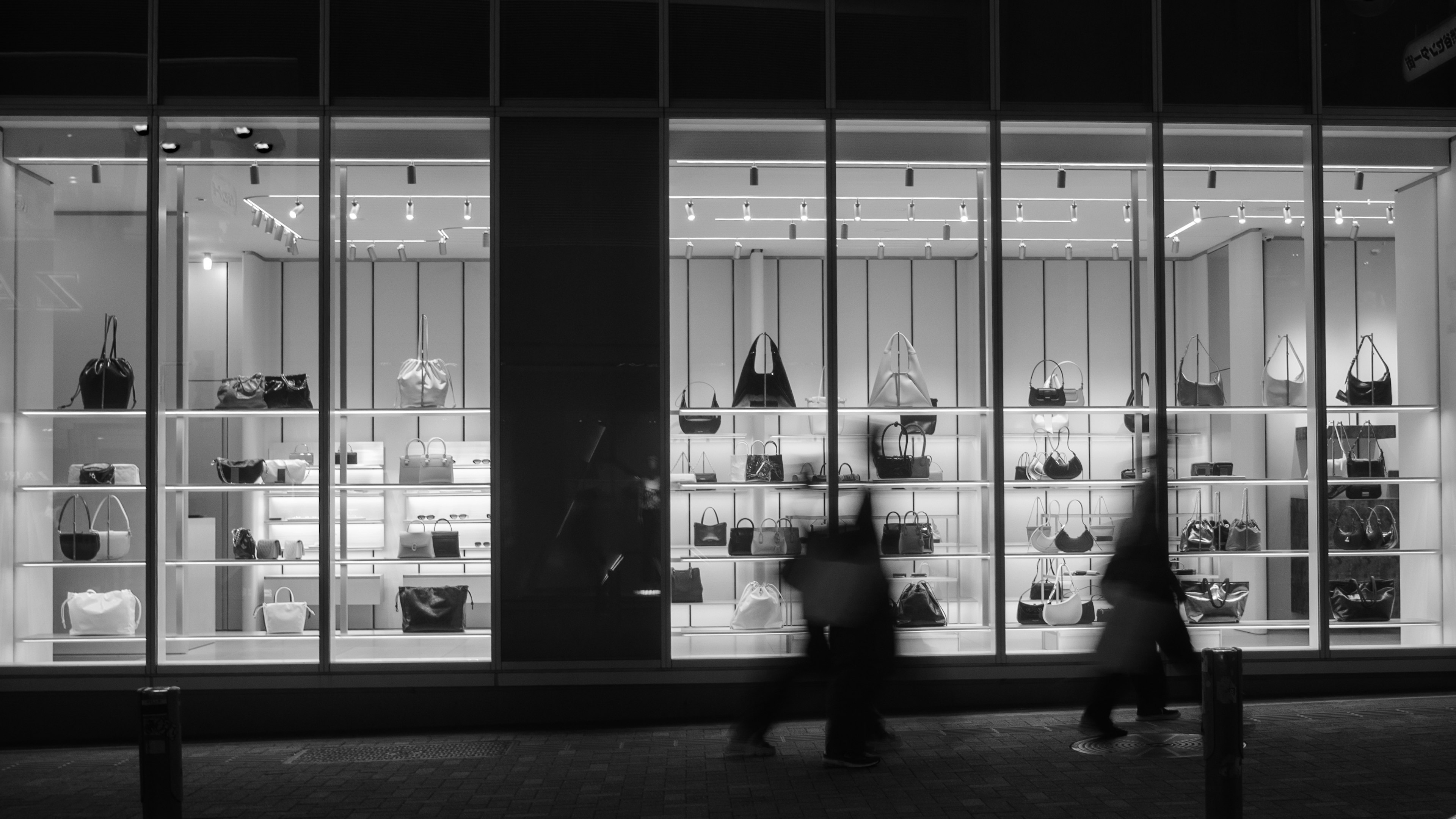 Nighttime storefront displaying various handbags and passing pedestrians