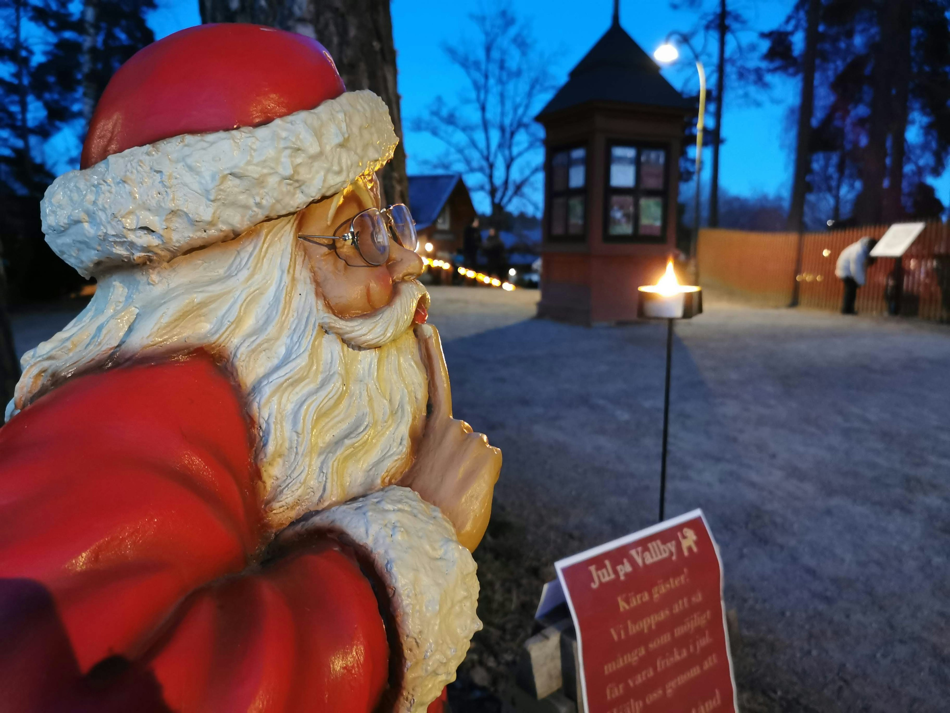 Santa Claus Statue mit Finger auf den Lippen in einer verschneiten Umgebung mit einer Hütte im Hintergrund