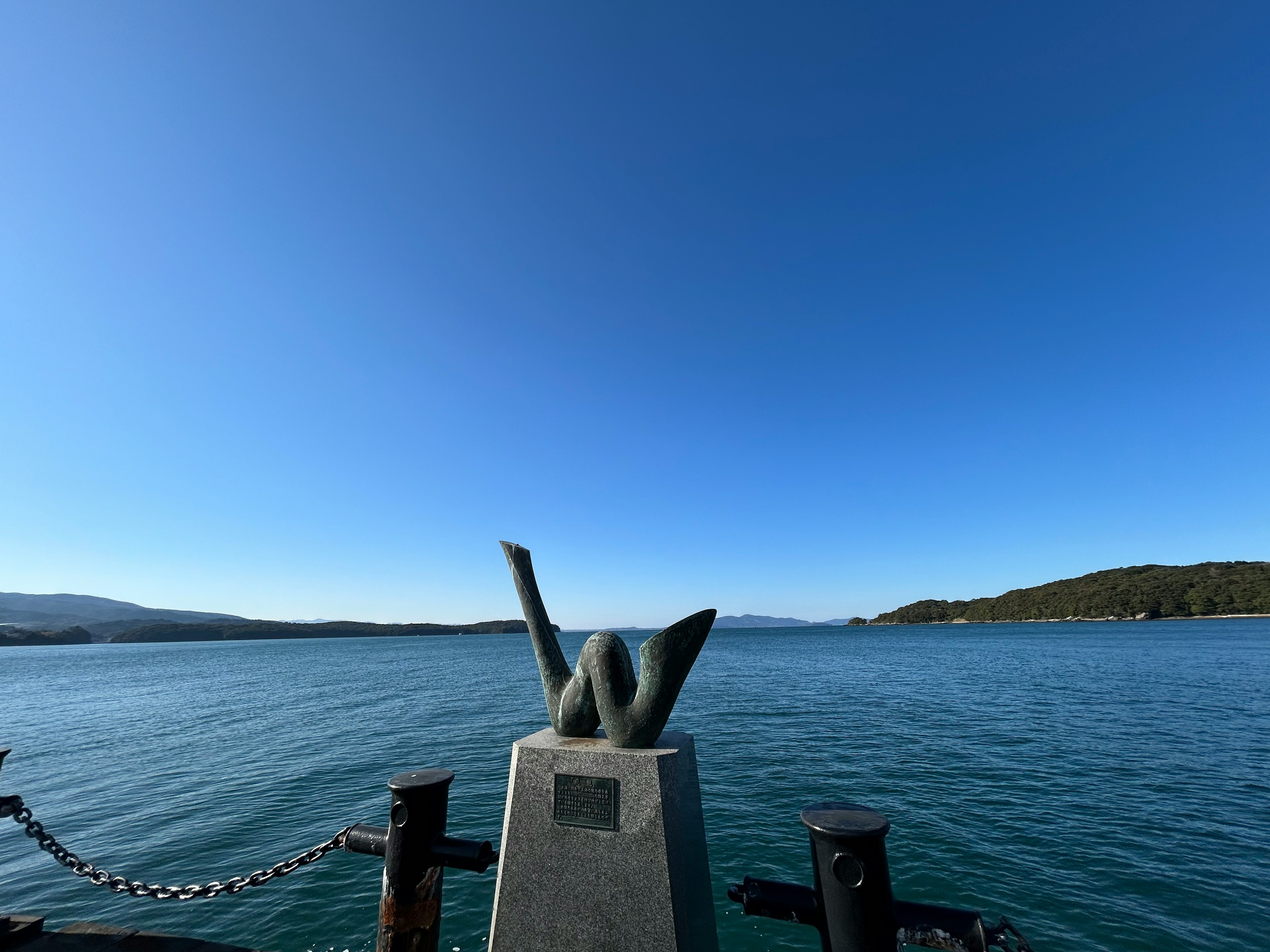 A monument with abstract sculpture against a clear blue sky and calm water