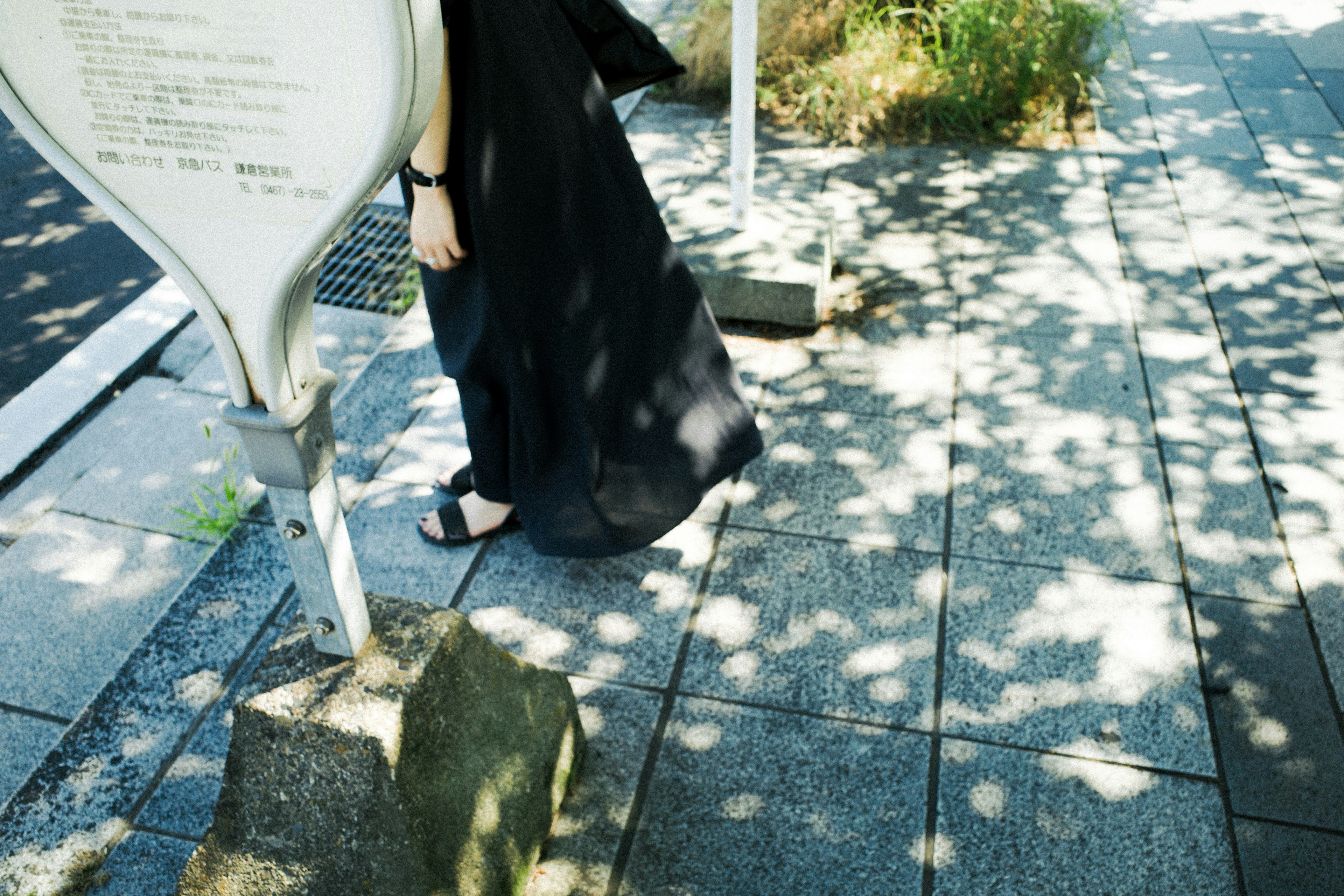 Image of a person in black clothing walking on a paved sidewalk with shadows and greenery