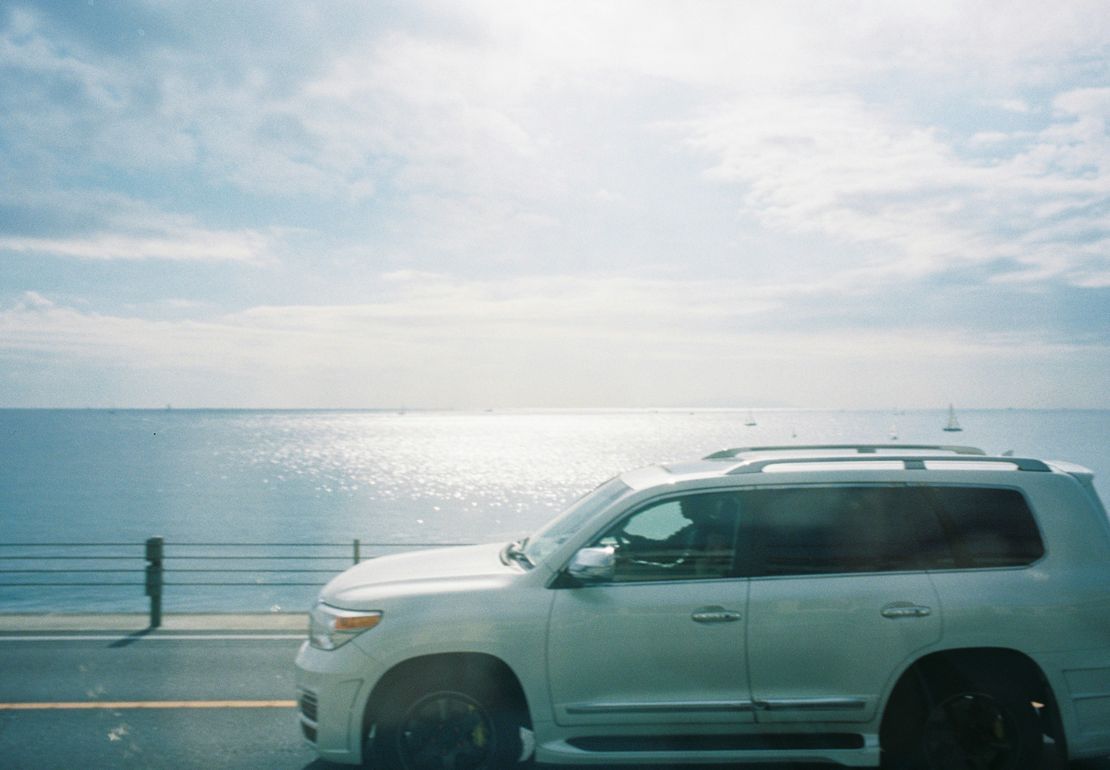 A white SUV driving along a road with a blue ocean and bright sky in the background