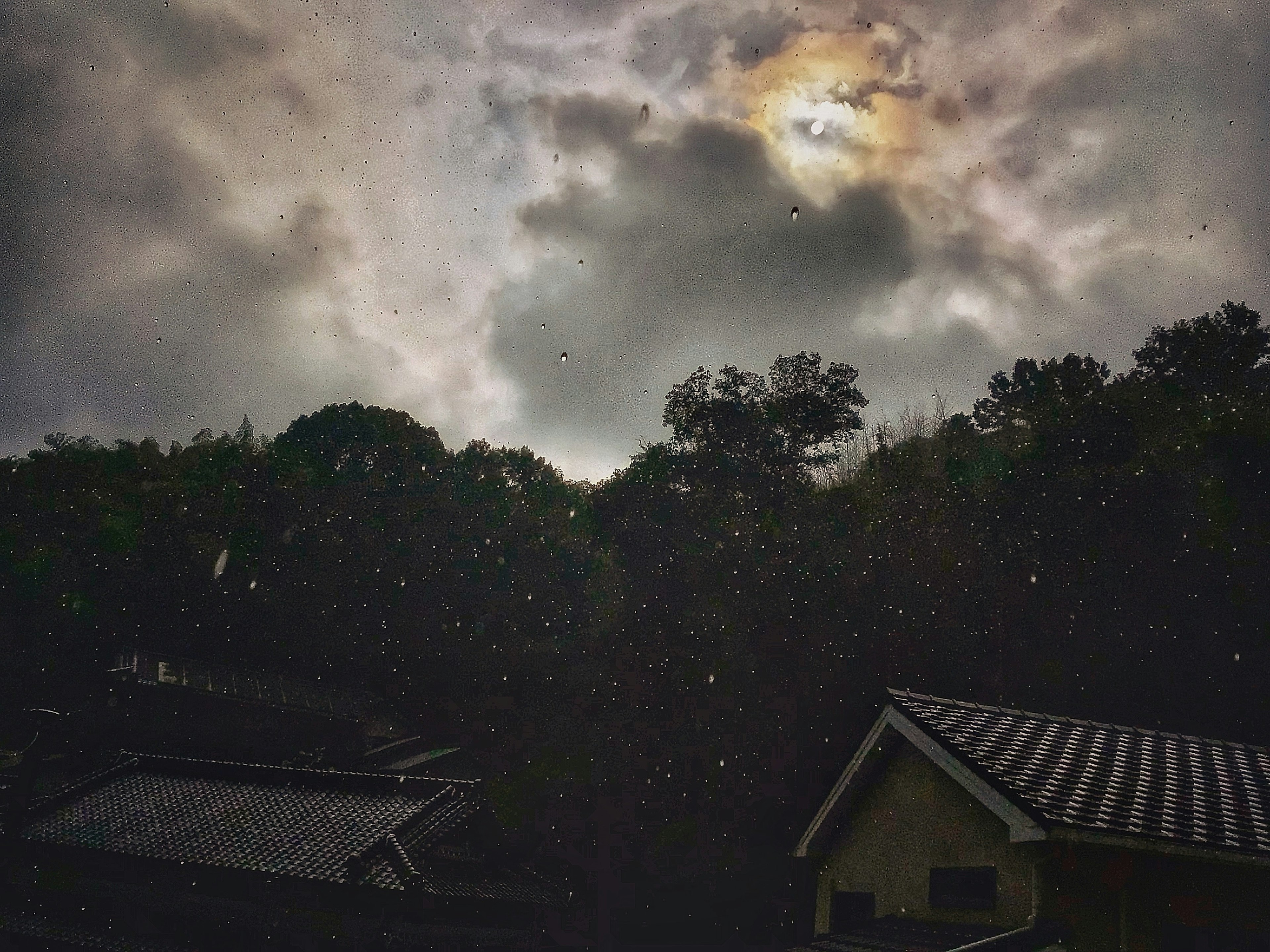 Langit gelap dengan awan dan hujan yang turun di atas rumah dengan bulan samar terlihat