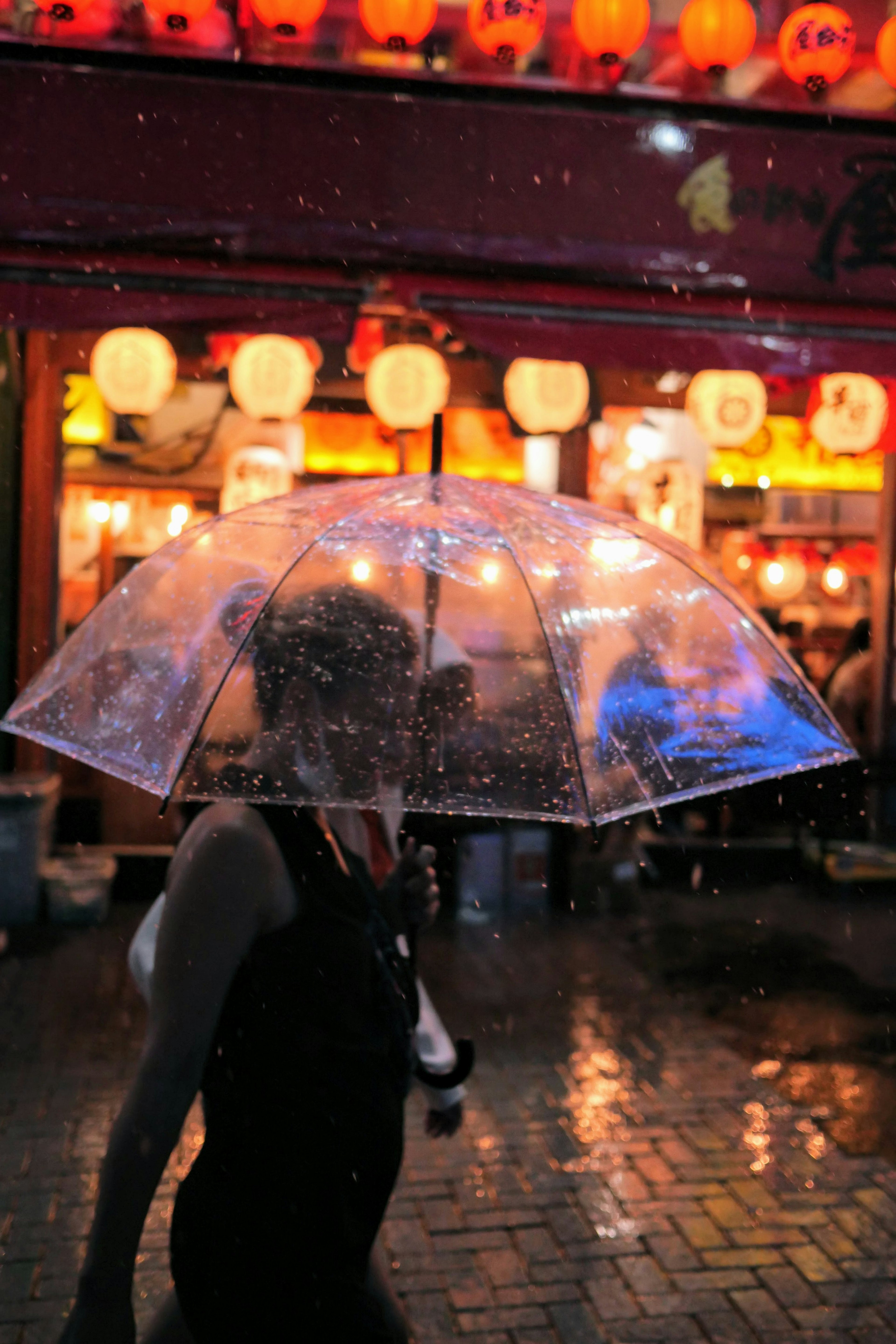 透明な傘を持つ女性が雨の中を歩いている街の風景 明るいランタンが照らす背景