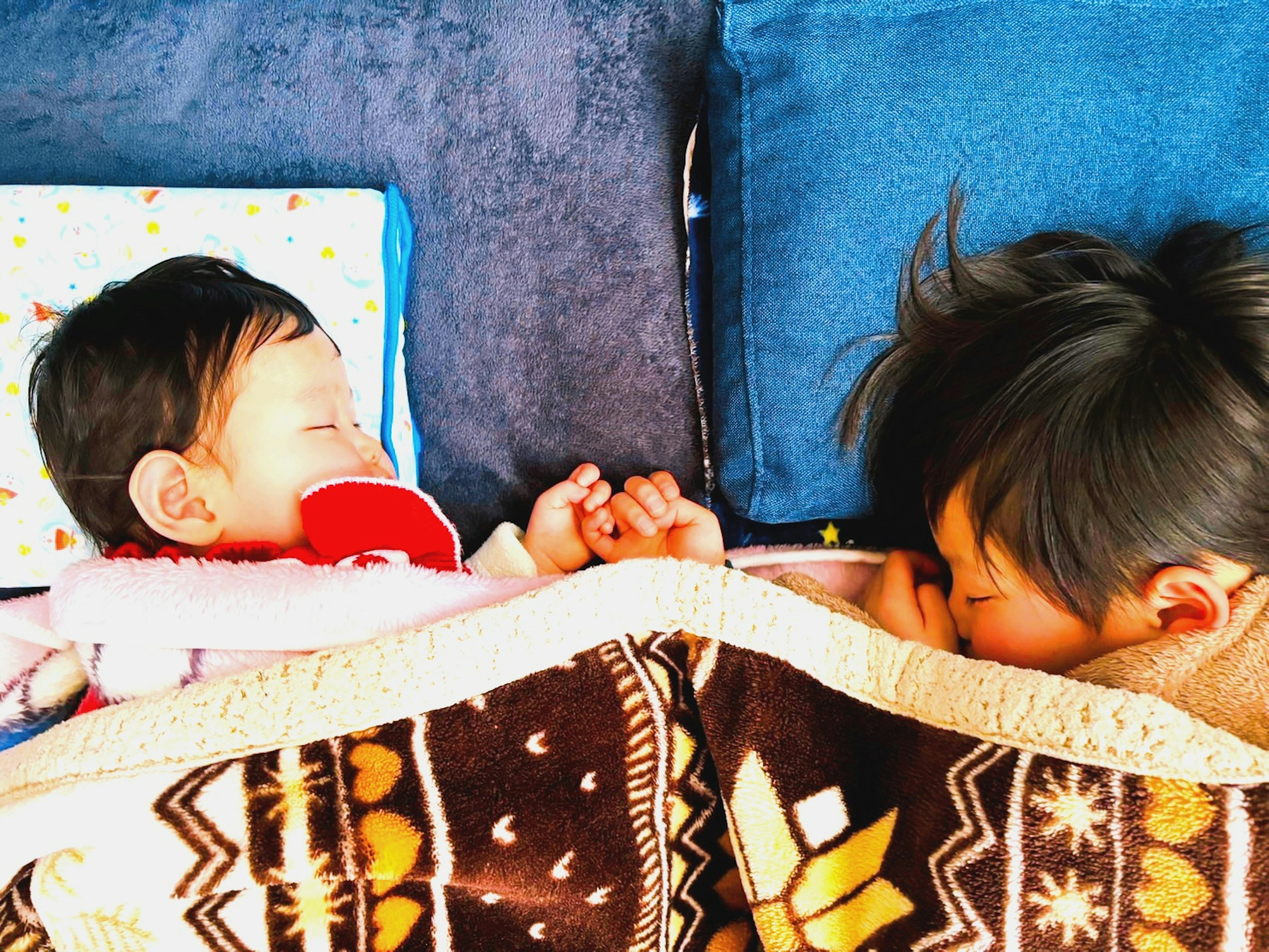 Dos niños durmiendo bajo una manta con almohadas azules al fondo