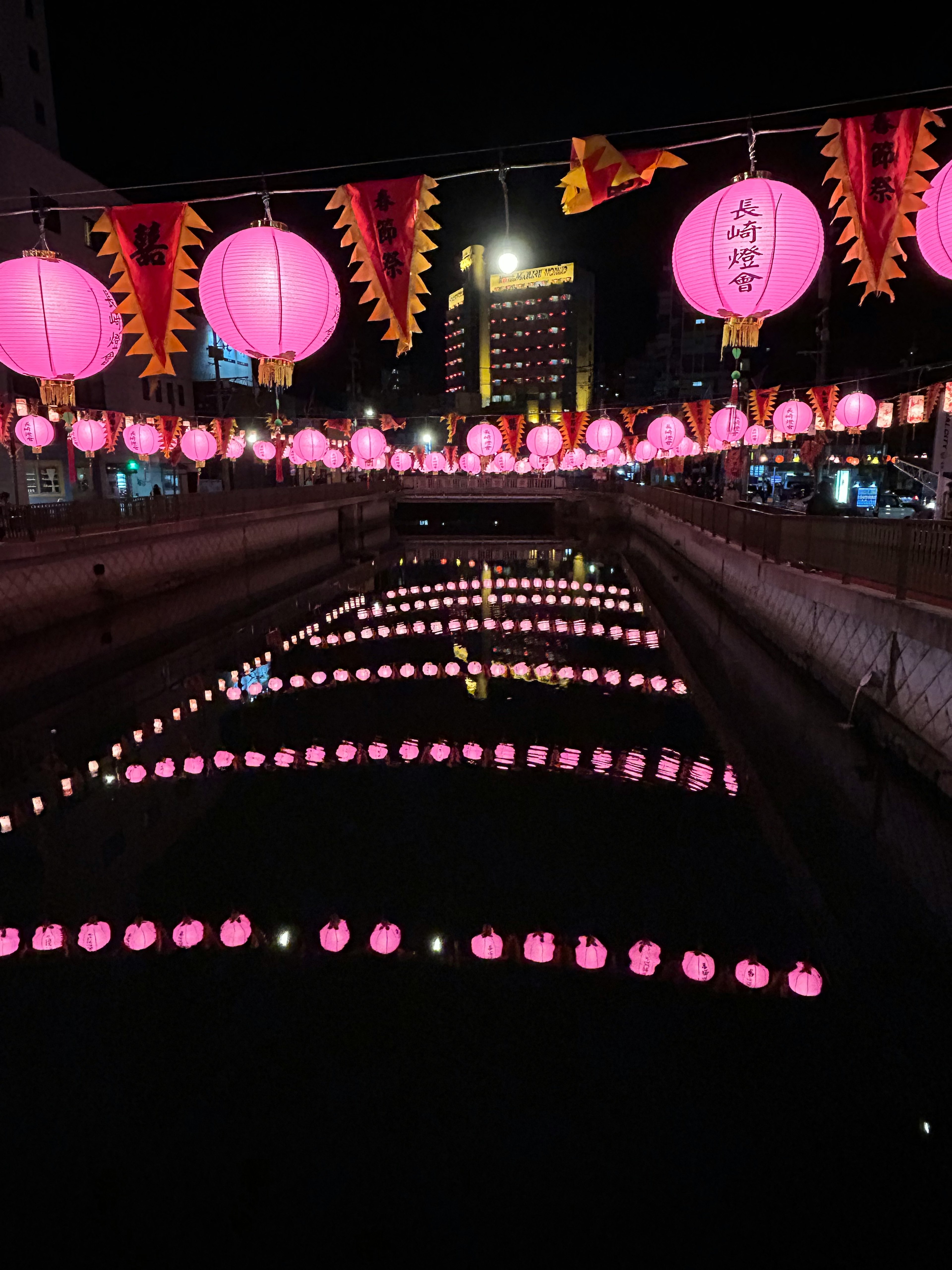 Filas de linternas rosas flotando en un río por la noche con luces de la ciudad