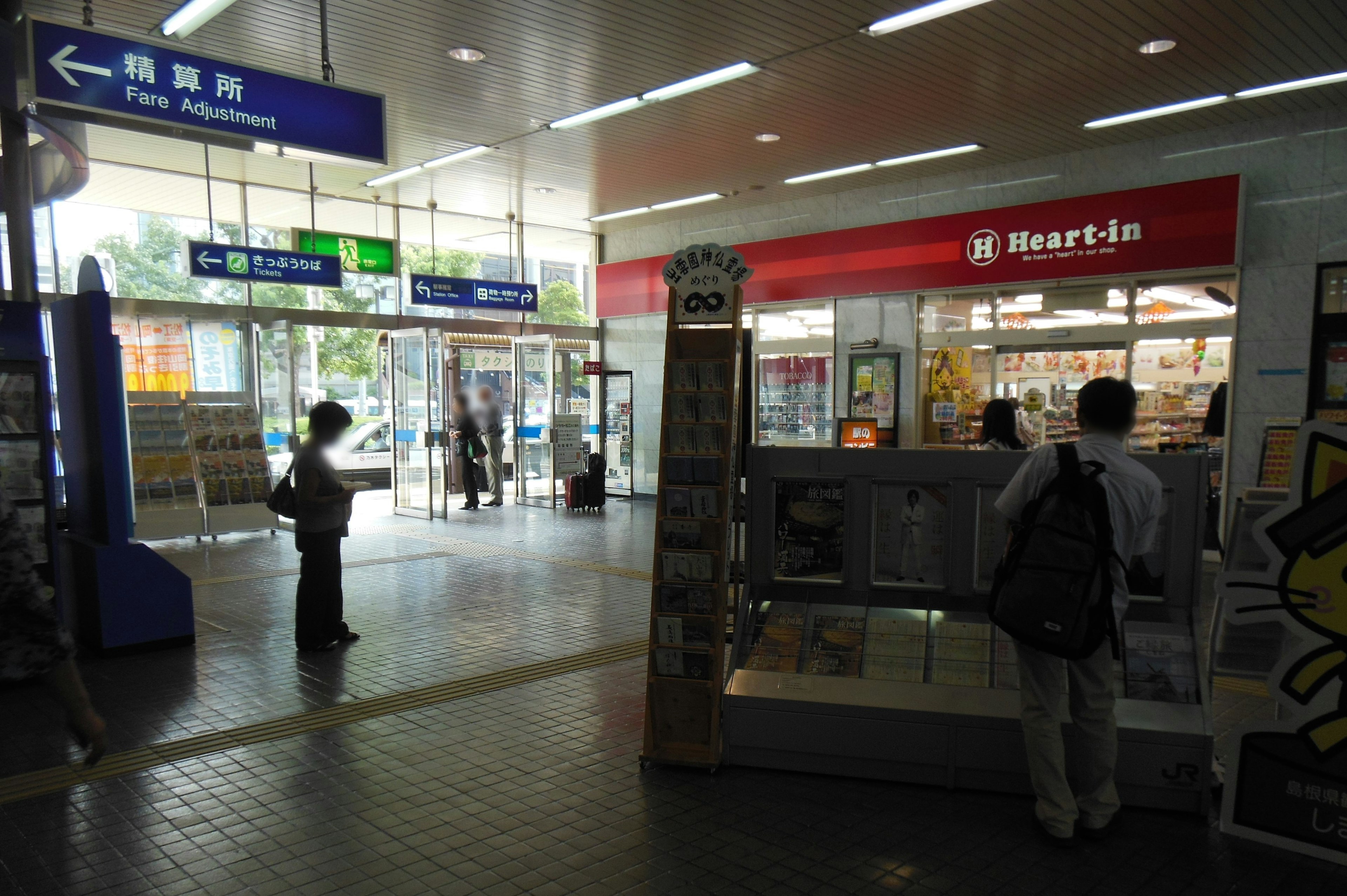 Interior stasiun kereta dengan tanda dan toko terlihat, pengunjung hadir