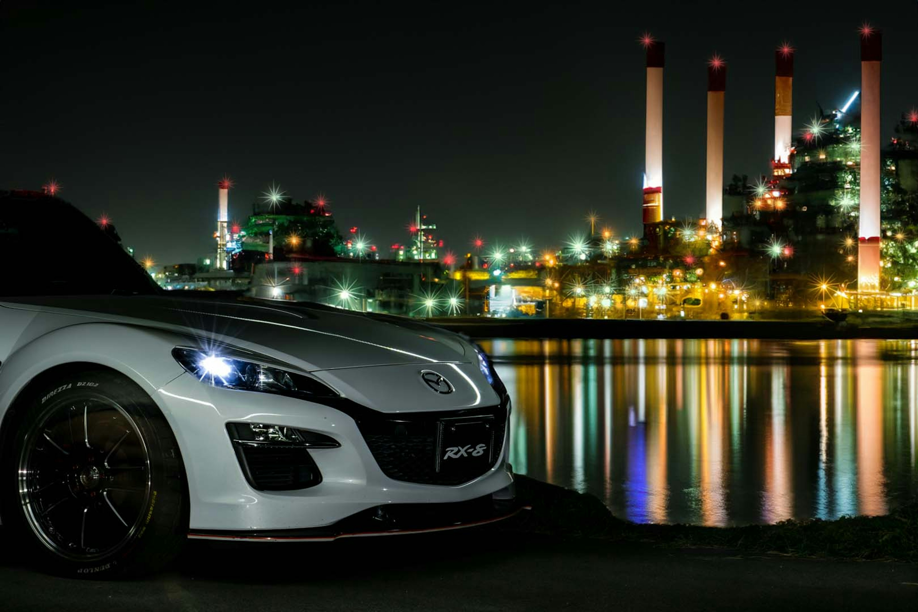 Close-up of a white sports car with a factory skyline at night