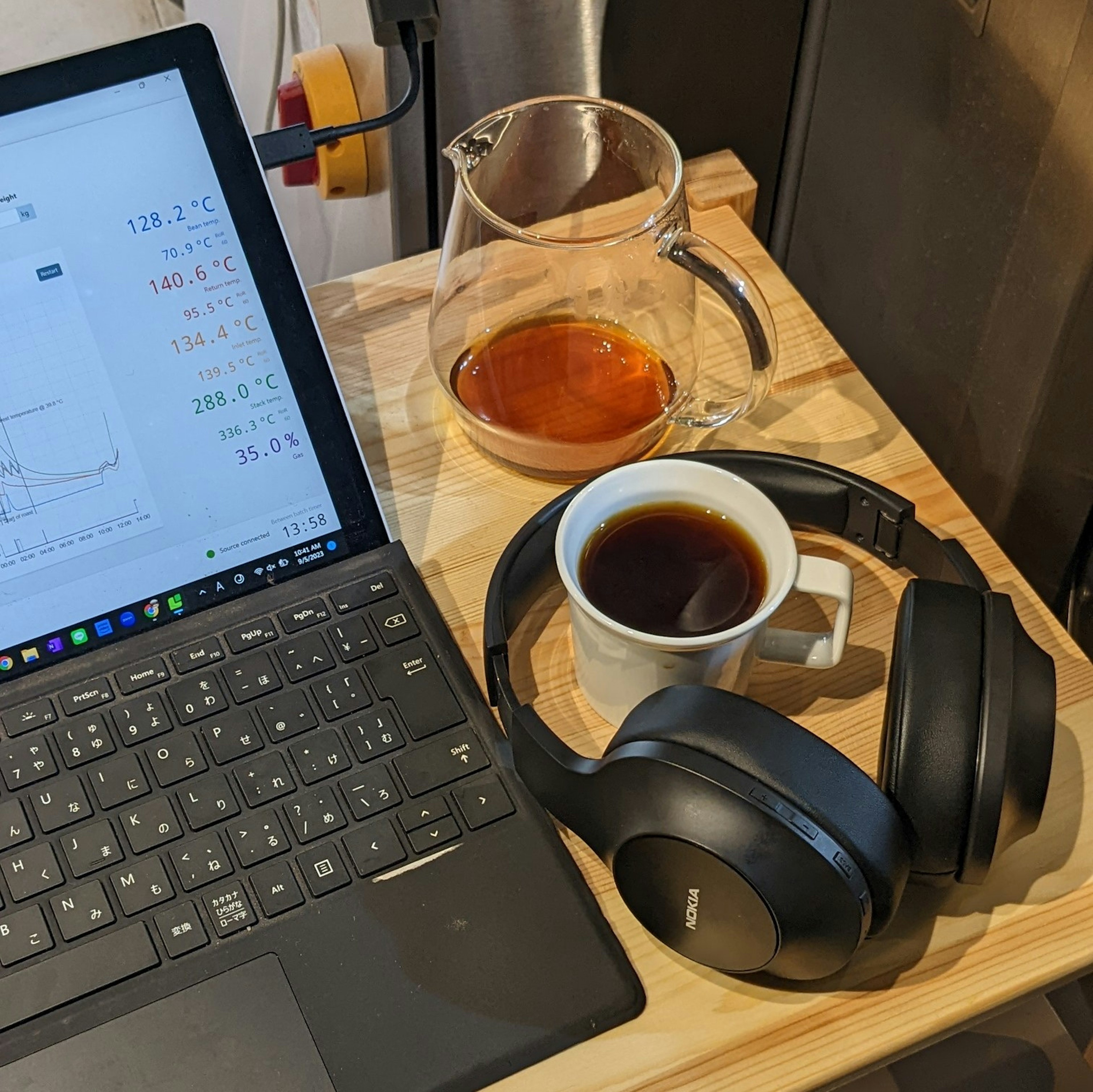 Laptop dengan spreadsheet, cangkir kopi, pot kopi, dan headphone di atas meja kayu