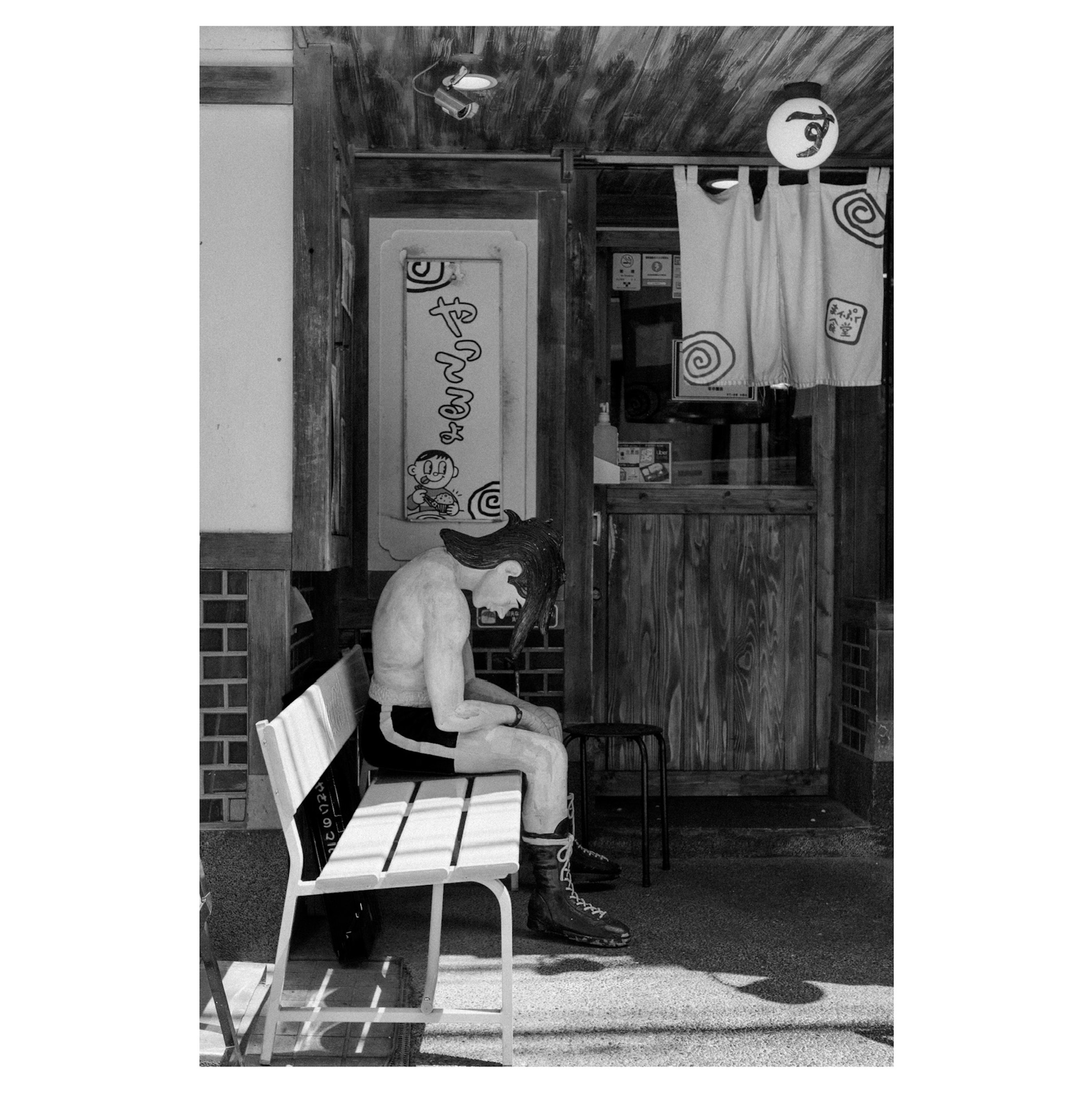 Black and white photo of a mannequin sitting on a bench with a small shop in the background featuring hanging curtains