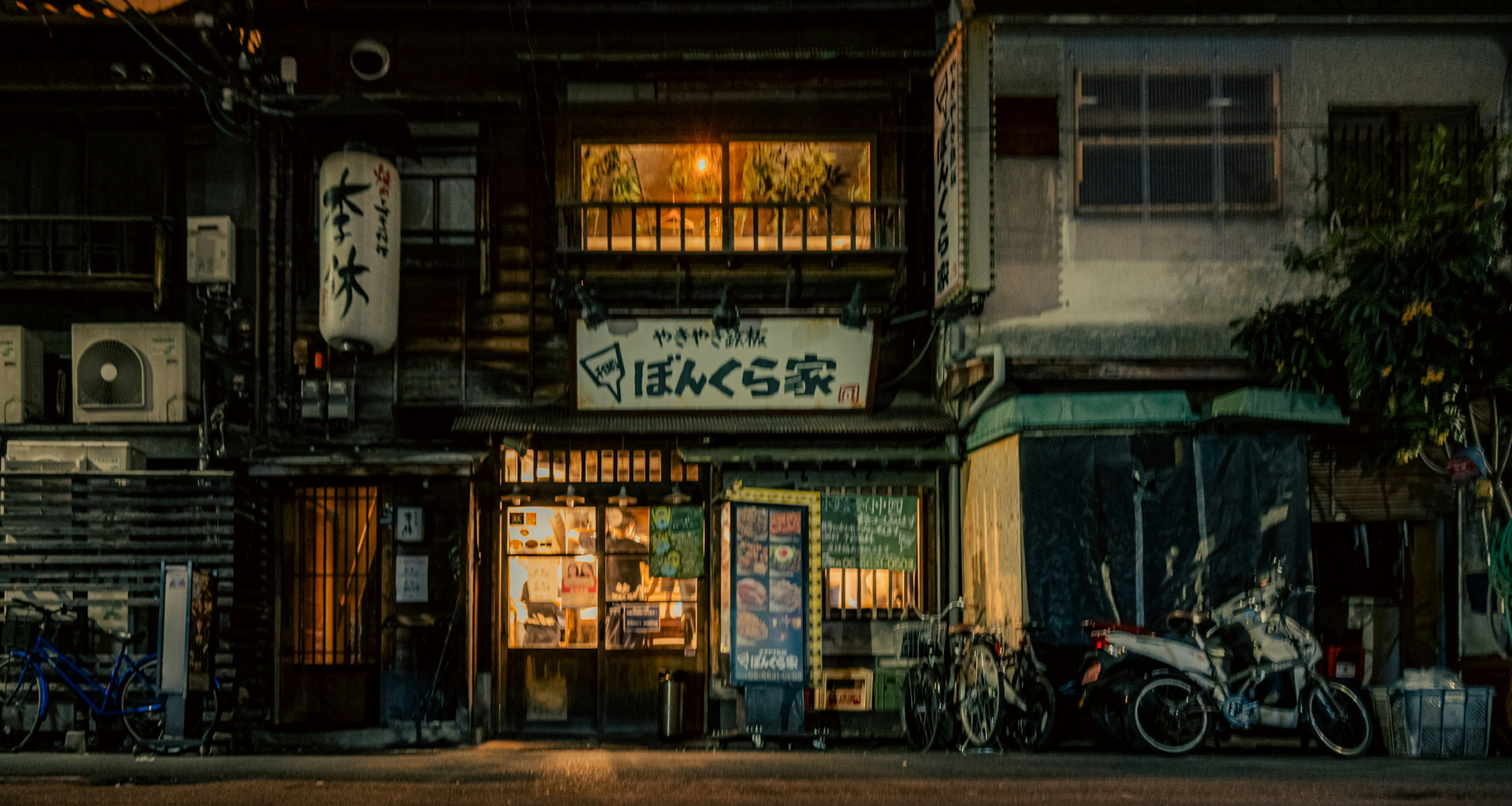 古い日本の商店街の夜景で、照明が温かく、バイクが停まっている