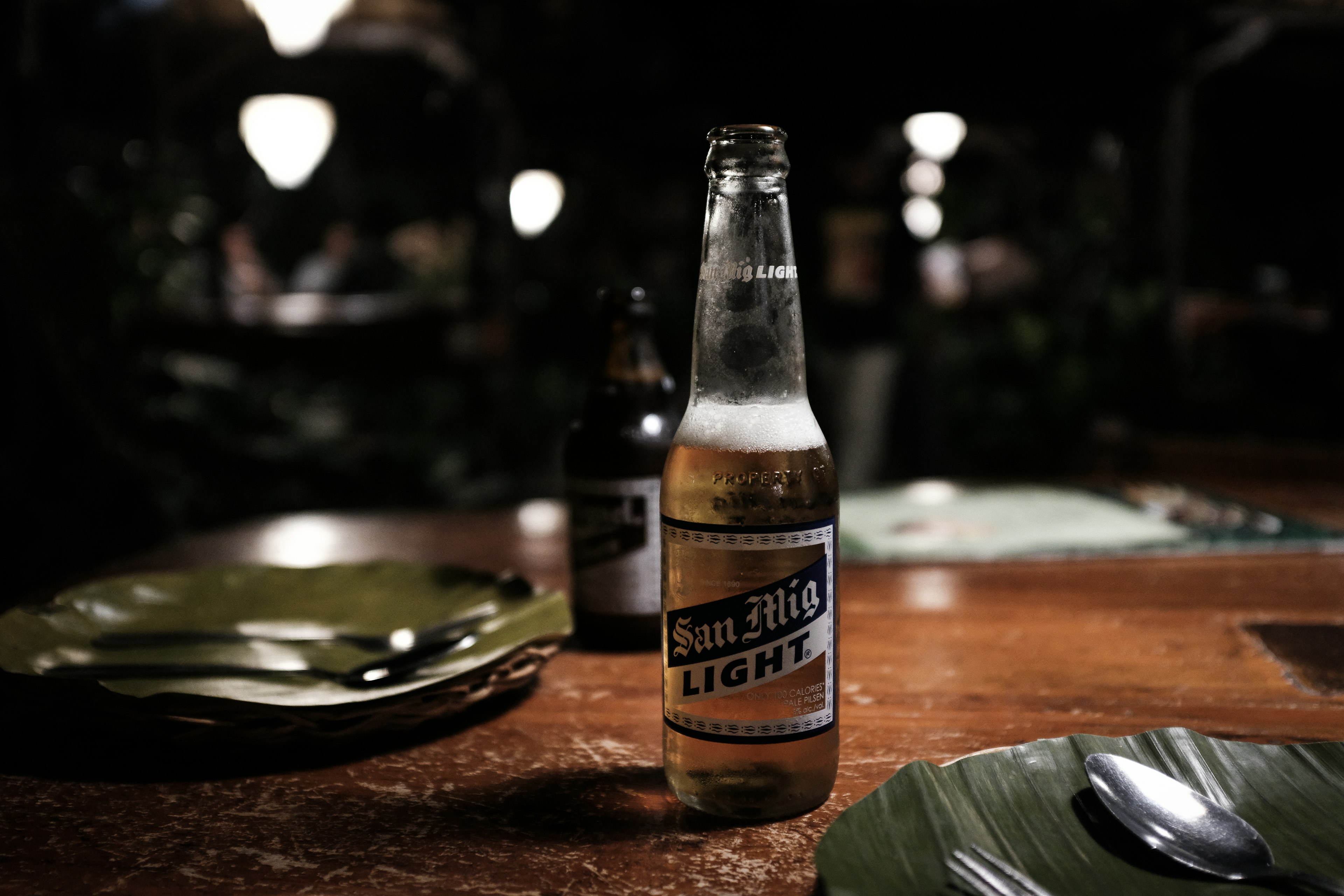 Une bouteille de bière San Miguel Light sur une table en bois avec des assiettes vertes dans un cadre faiblement éclairé