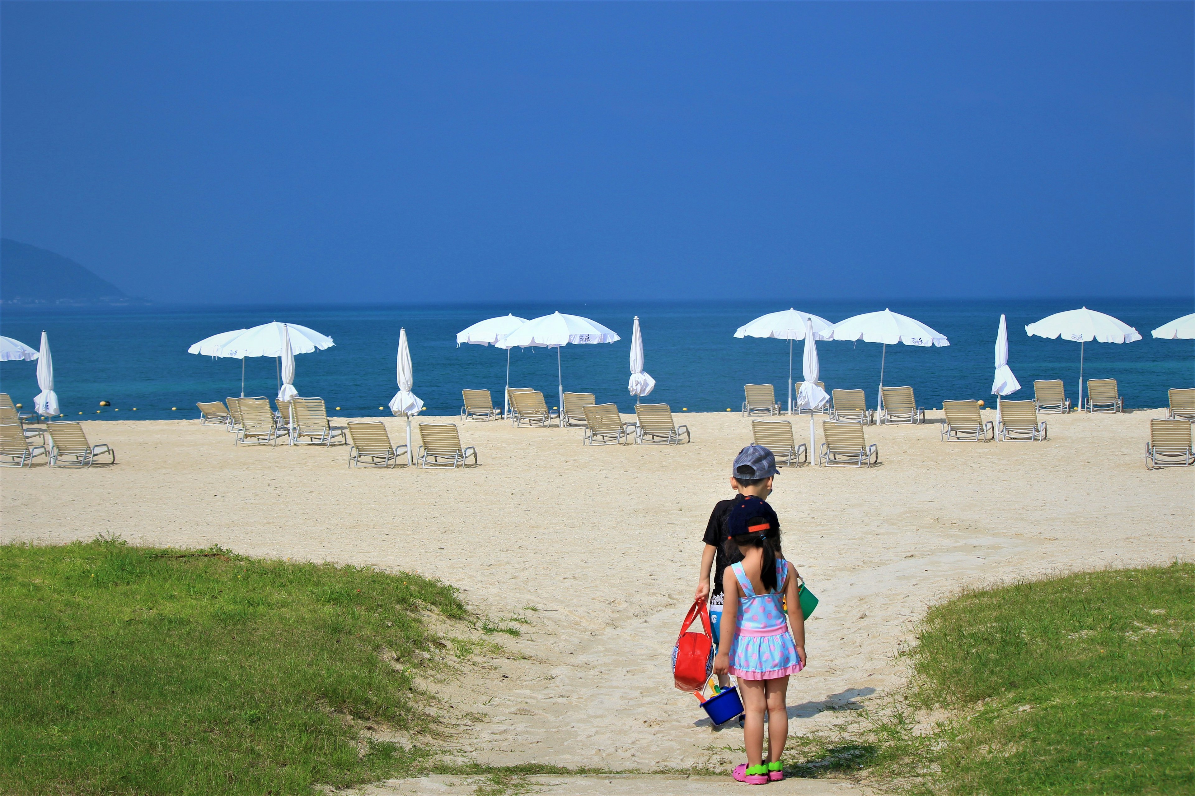 Anak-anak berjalan menuju pantai dengan payung putih dan pasir