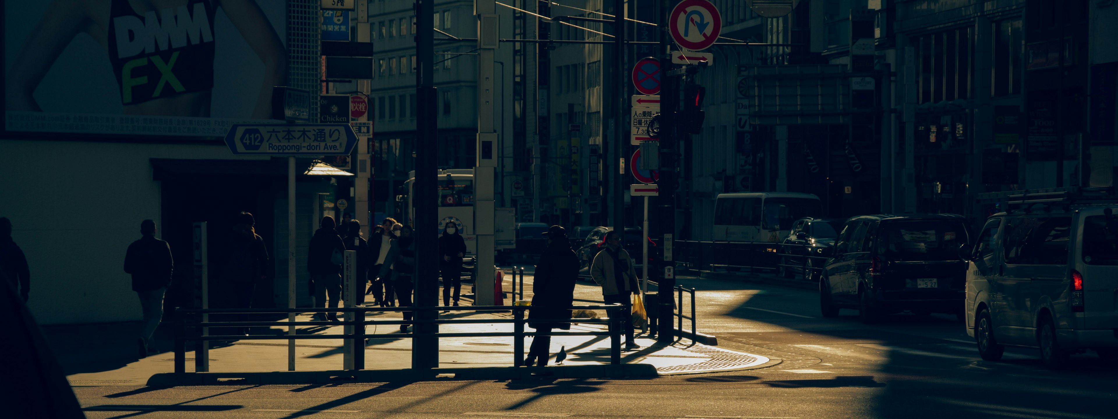 People standing at a dark street corner with passing cars during dusk