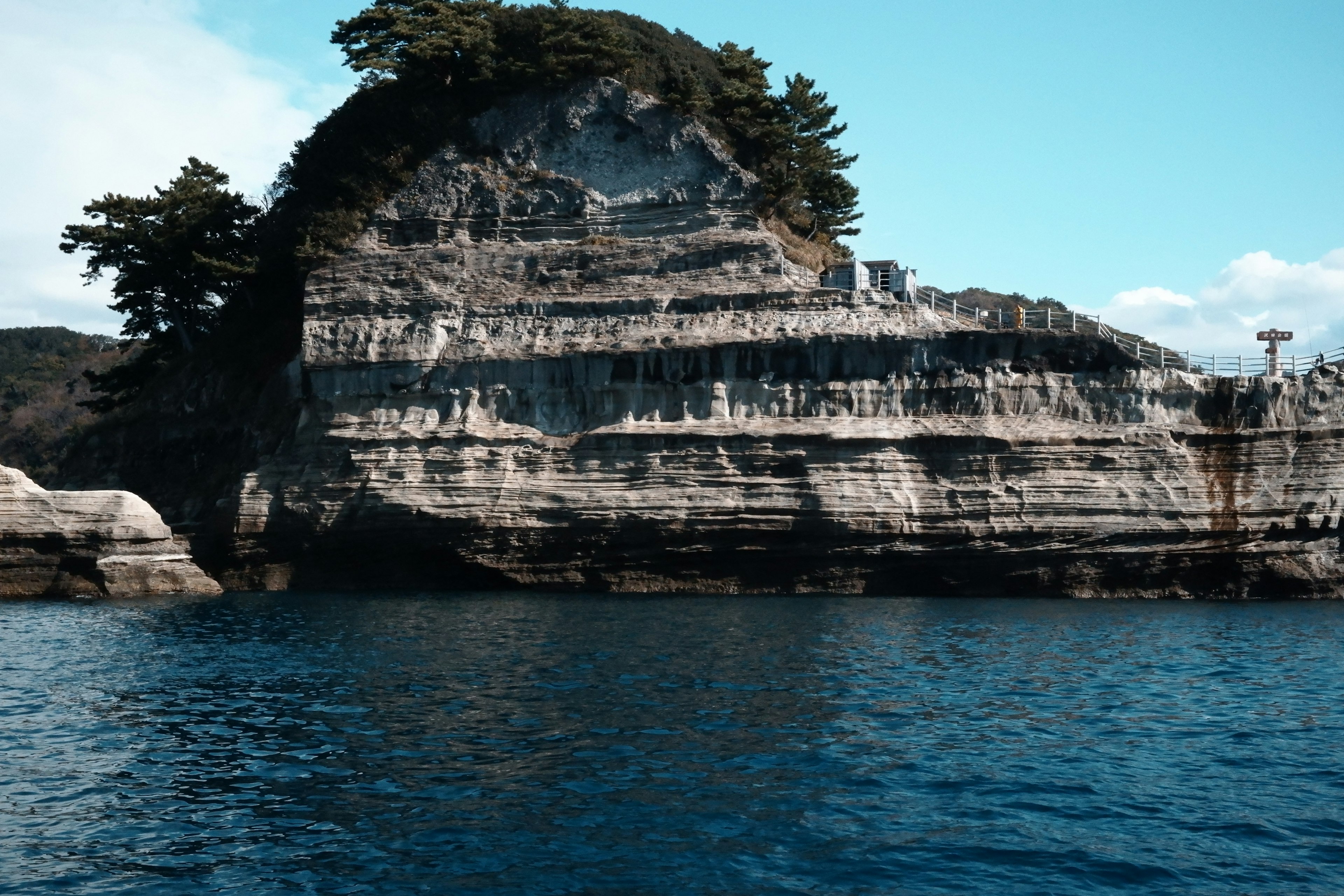 Rocky cliff with layered sedimentary rock and pine trees near blue water