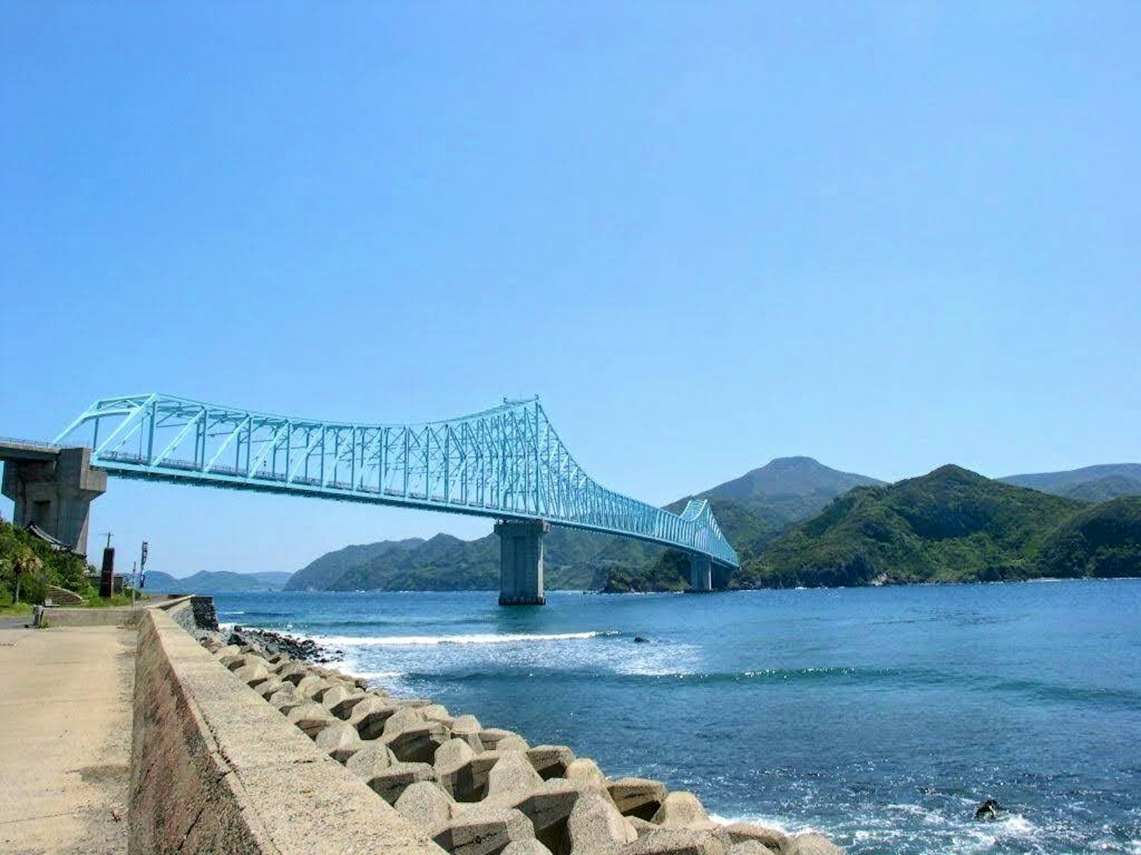 A blue bridge spans over the sea under a clear blue sky