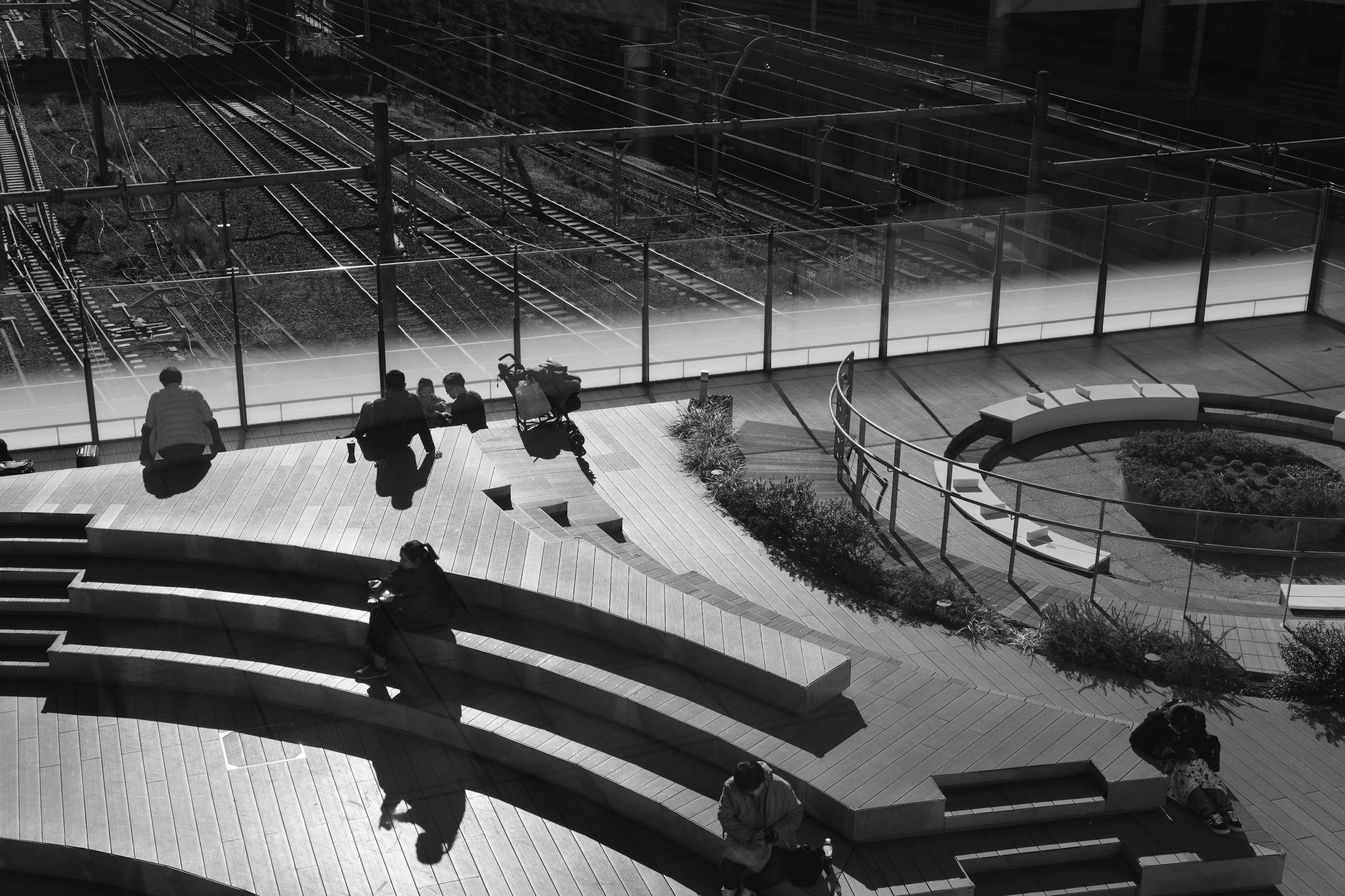 公園のベンチで座っている人々と背景の線路が見えるモノクロ写真