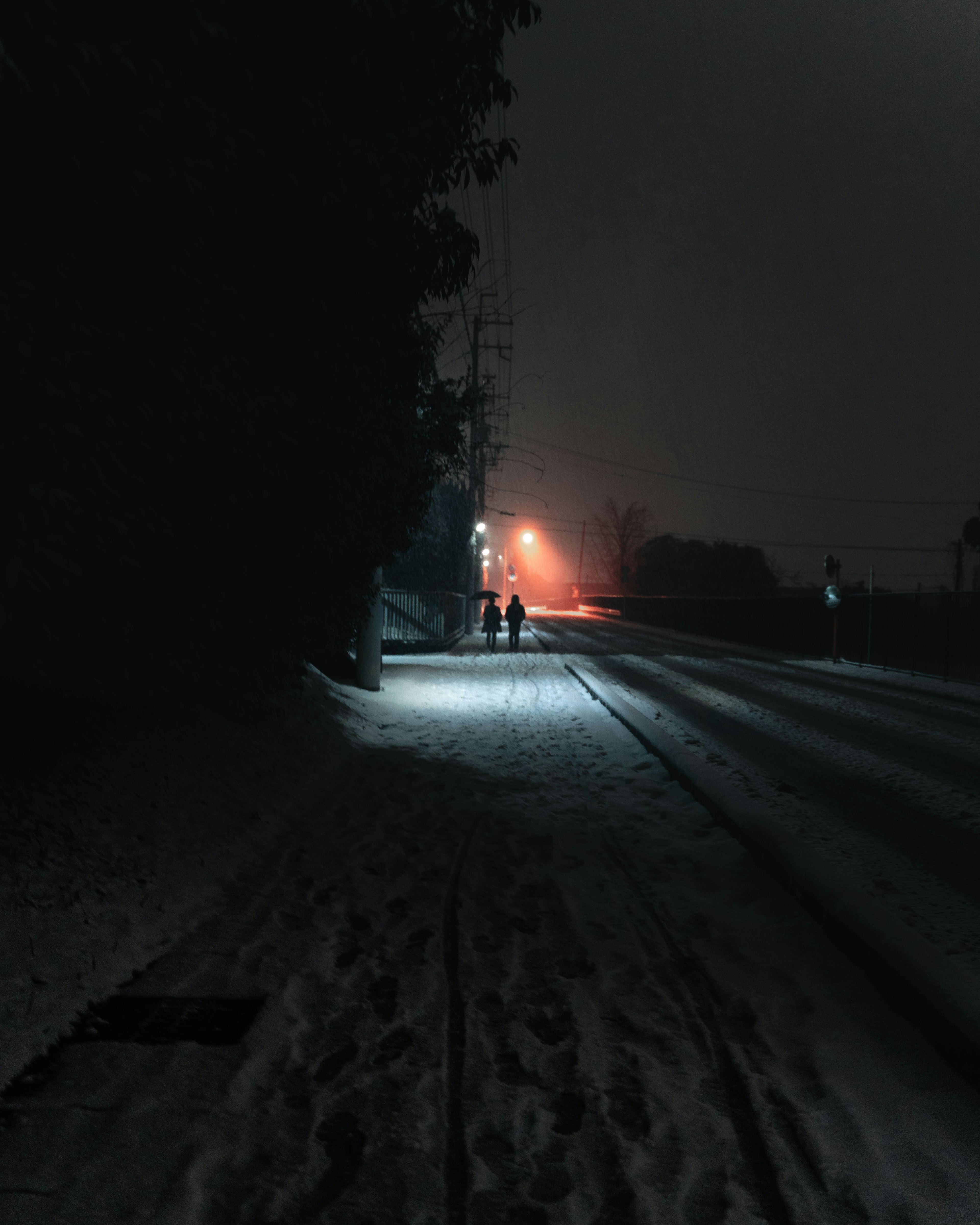 Two silhouettes walking on a snow-covered road with dim streetlights