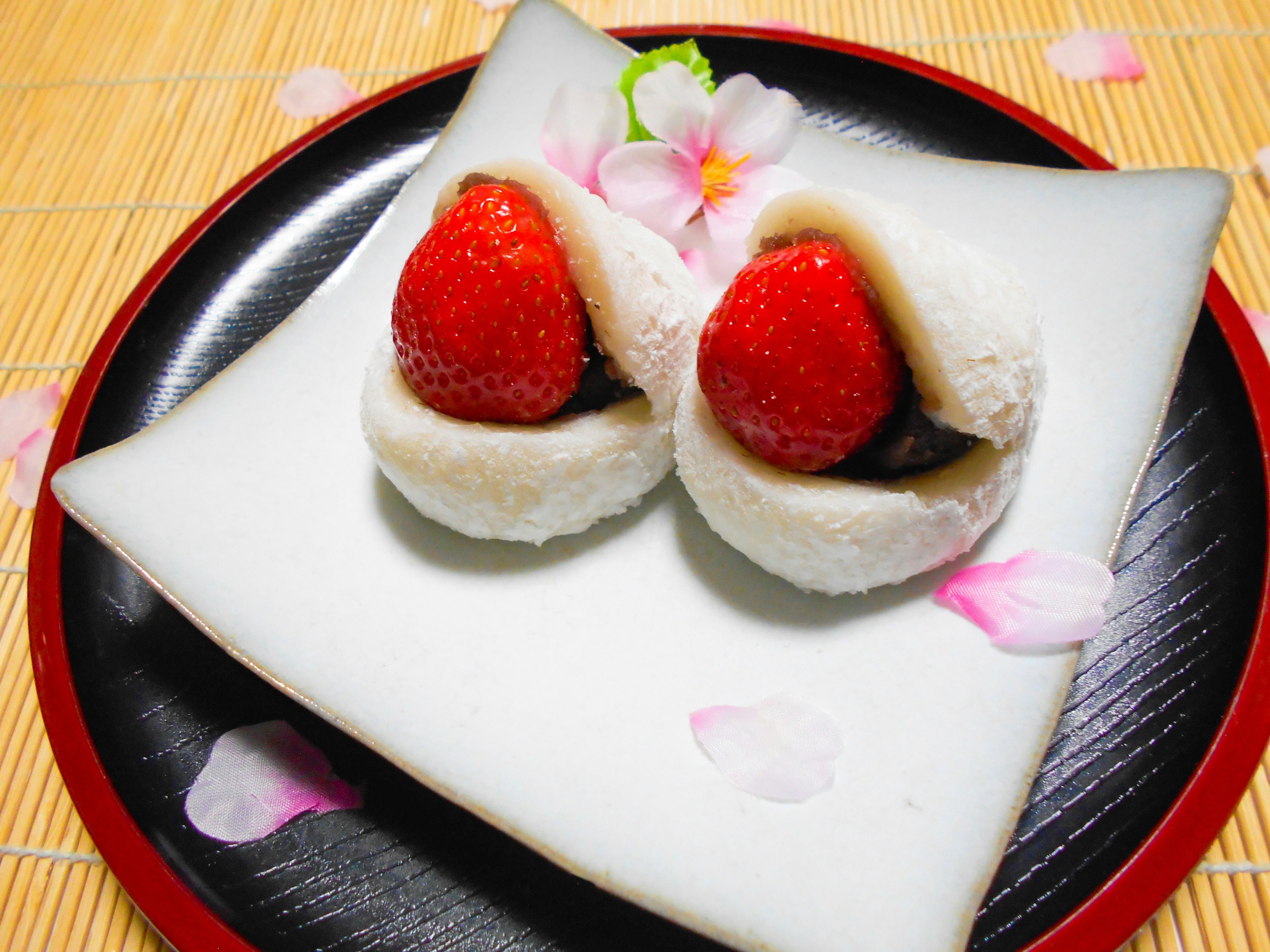 Two strawberry daifuku placed on a white plate with flower petals scattered around