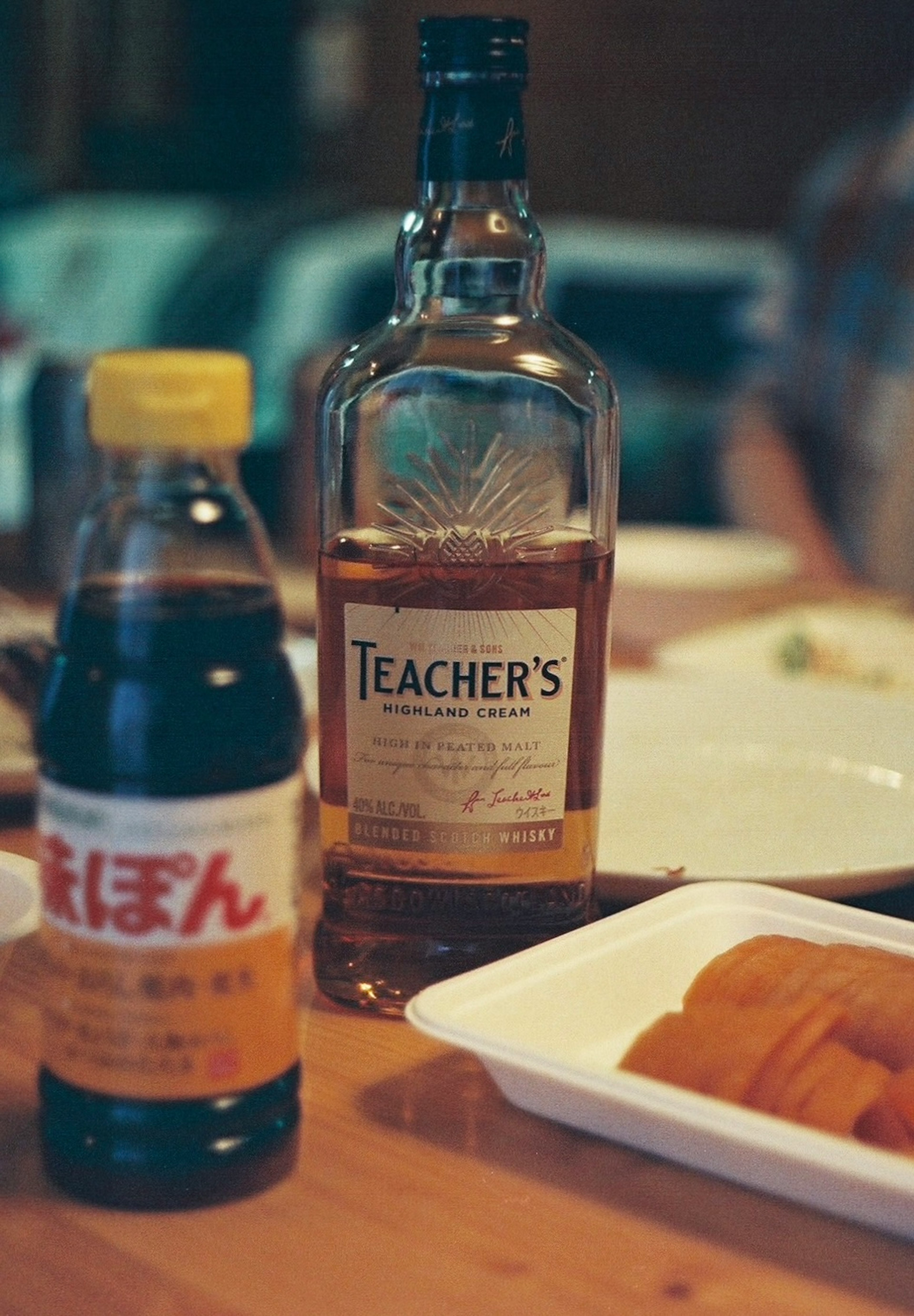 A whiskey bottle labeled Teacher's and a soy sauce bottle on a wooden table with food