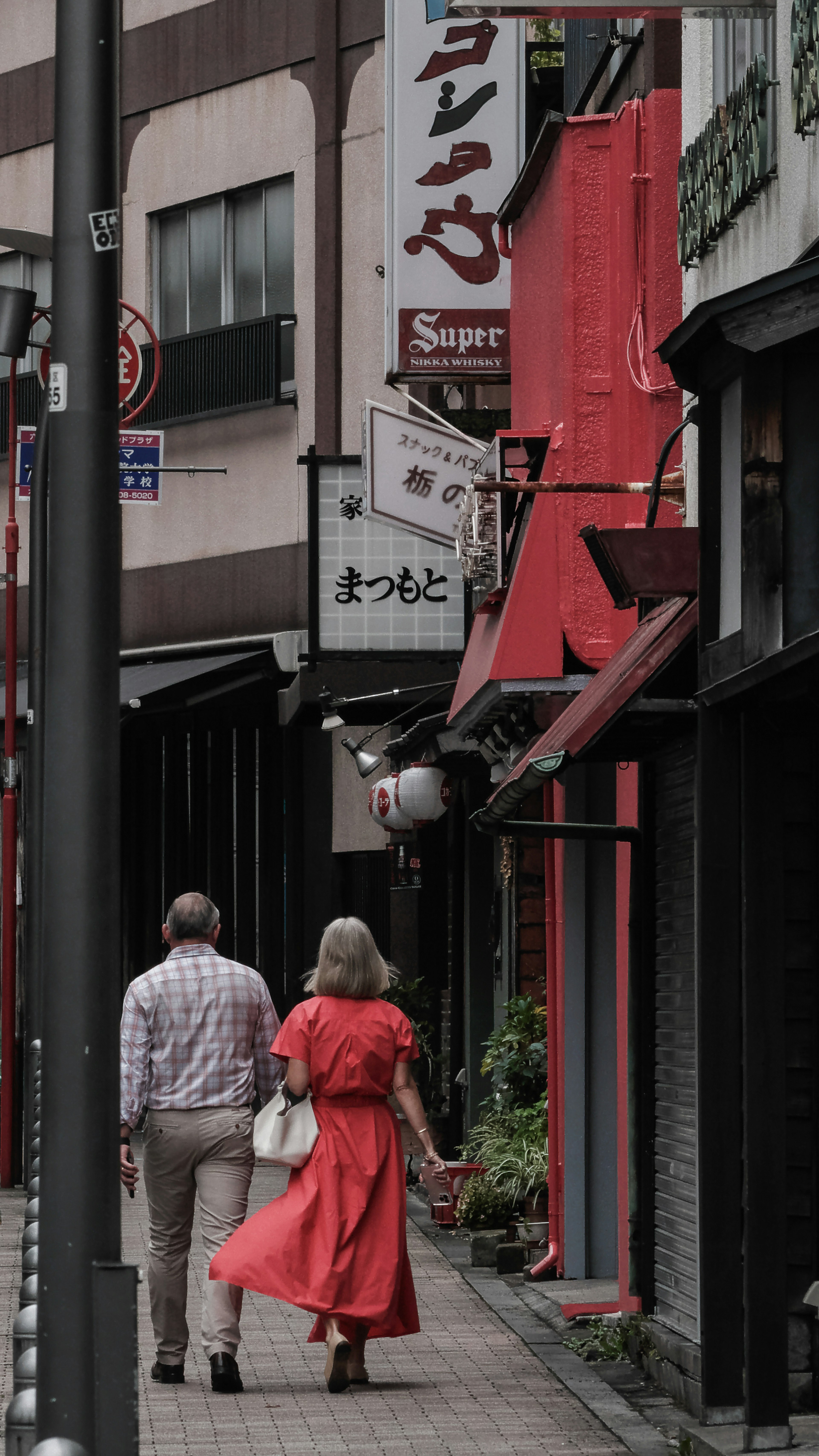 Une femme en robe rouge et un homme en chemise grise marchent dans une rue étroite bordée de magasins