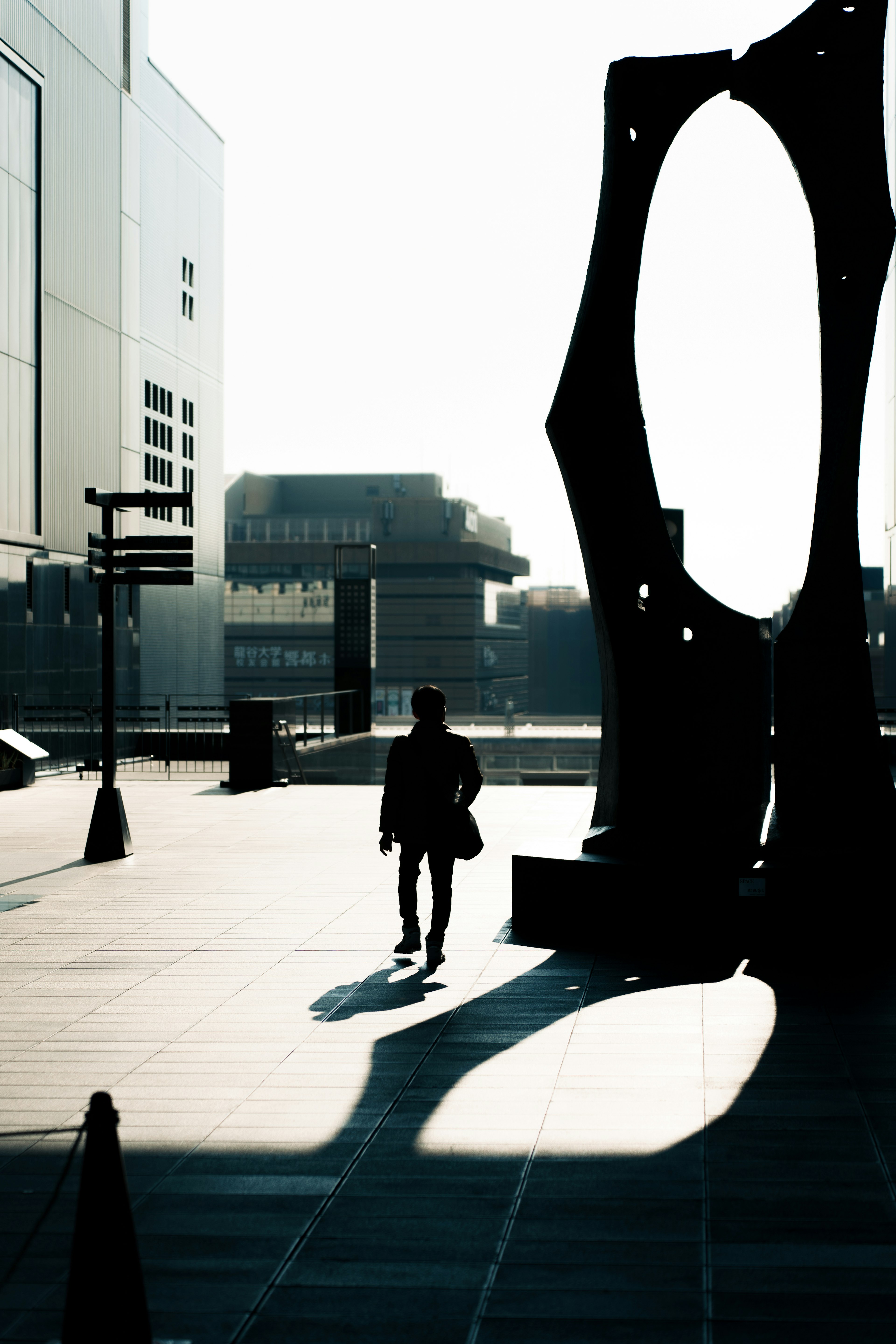 Silhouette of a person walking beside a sculpture in a sunlit urban setting