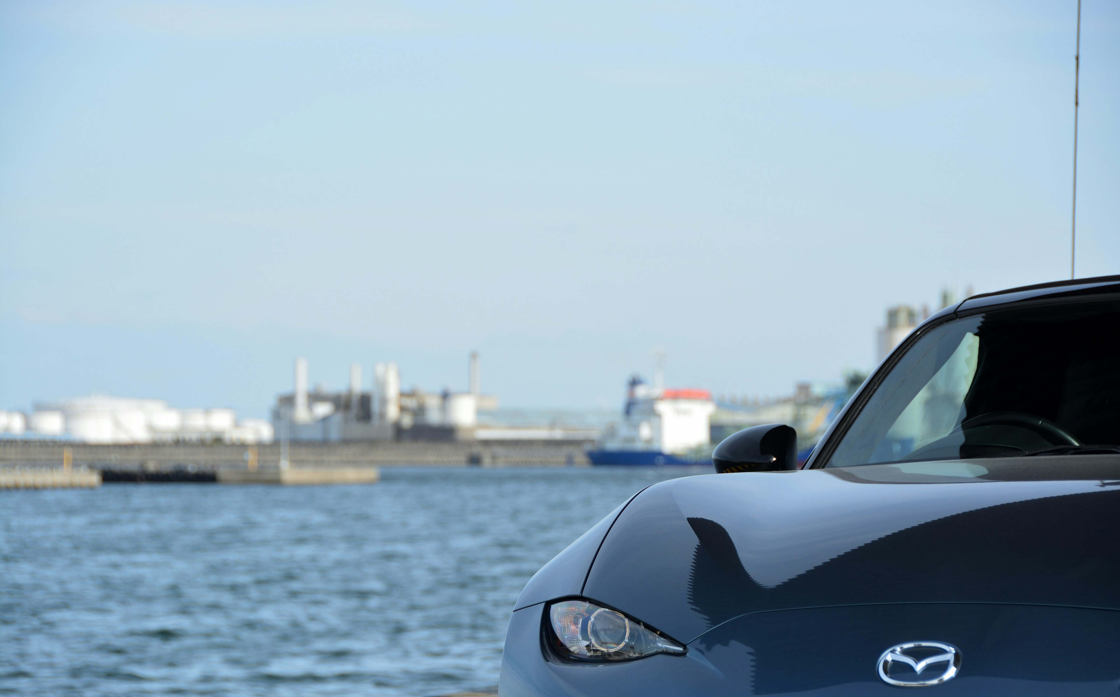 Front view of a black Mazda car near a harbor with industrial buildings in the background