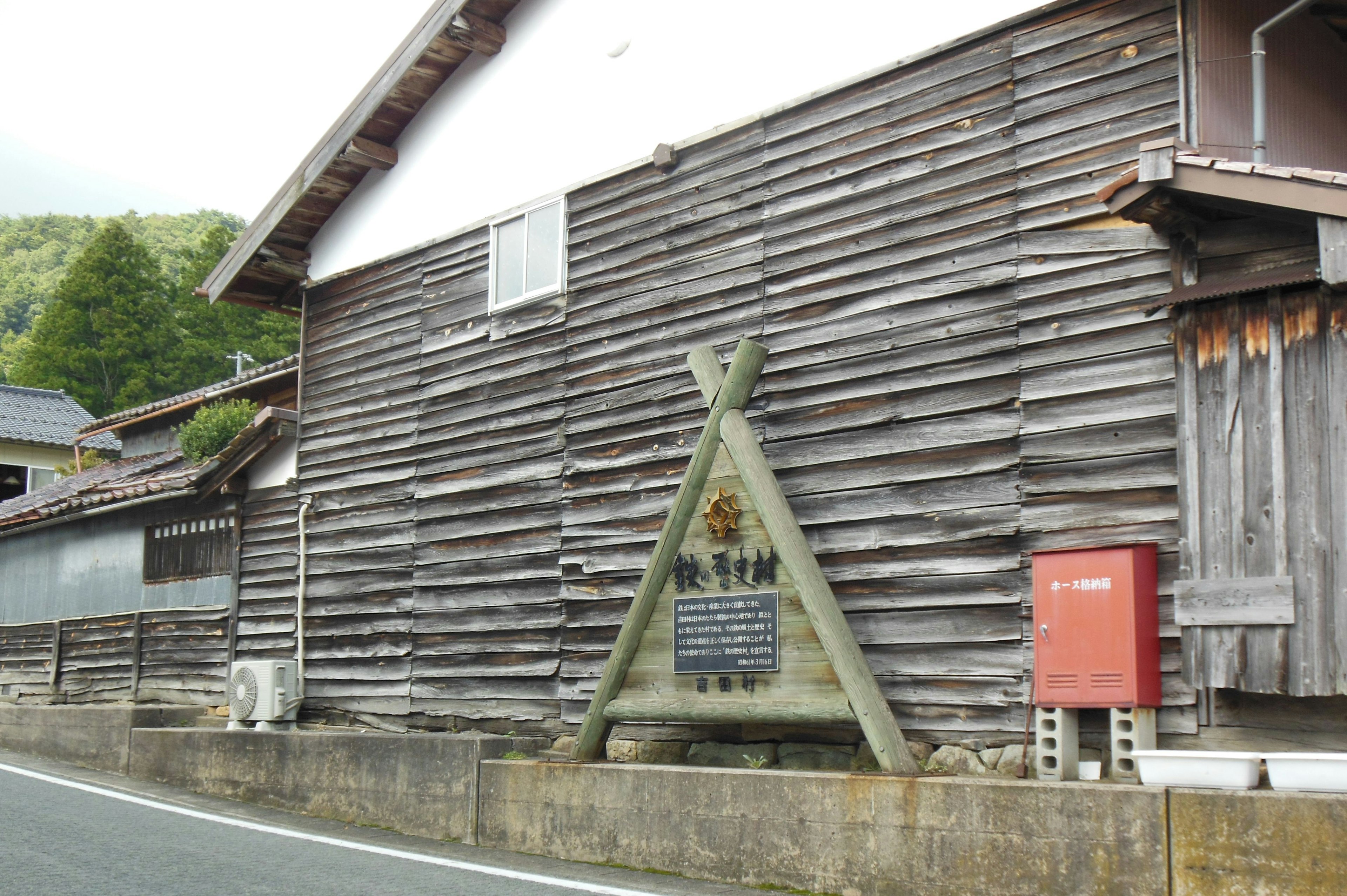 Außenansicht eines alten Holzhauses mit einem dreieckigen Schild und einem roten Briefkasten