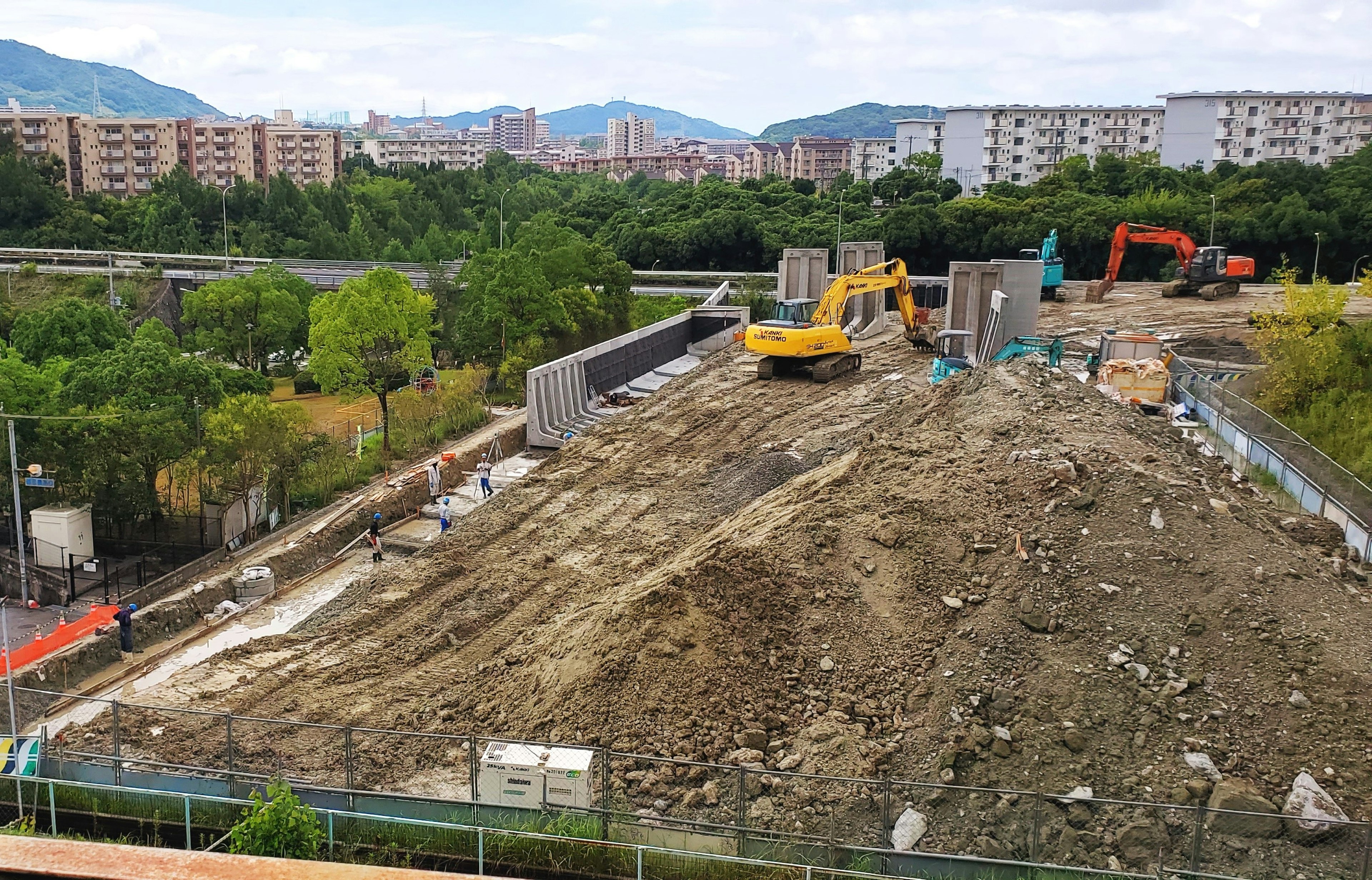 工事現場の風景、黄色い重機と土の山、周囲の建物、緑の木々