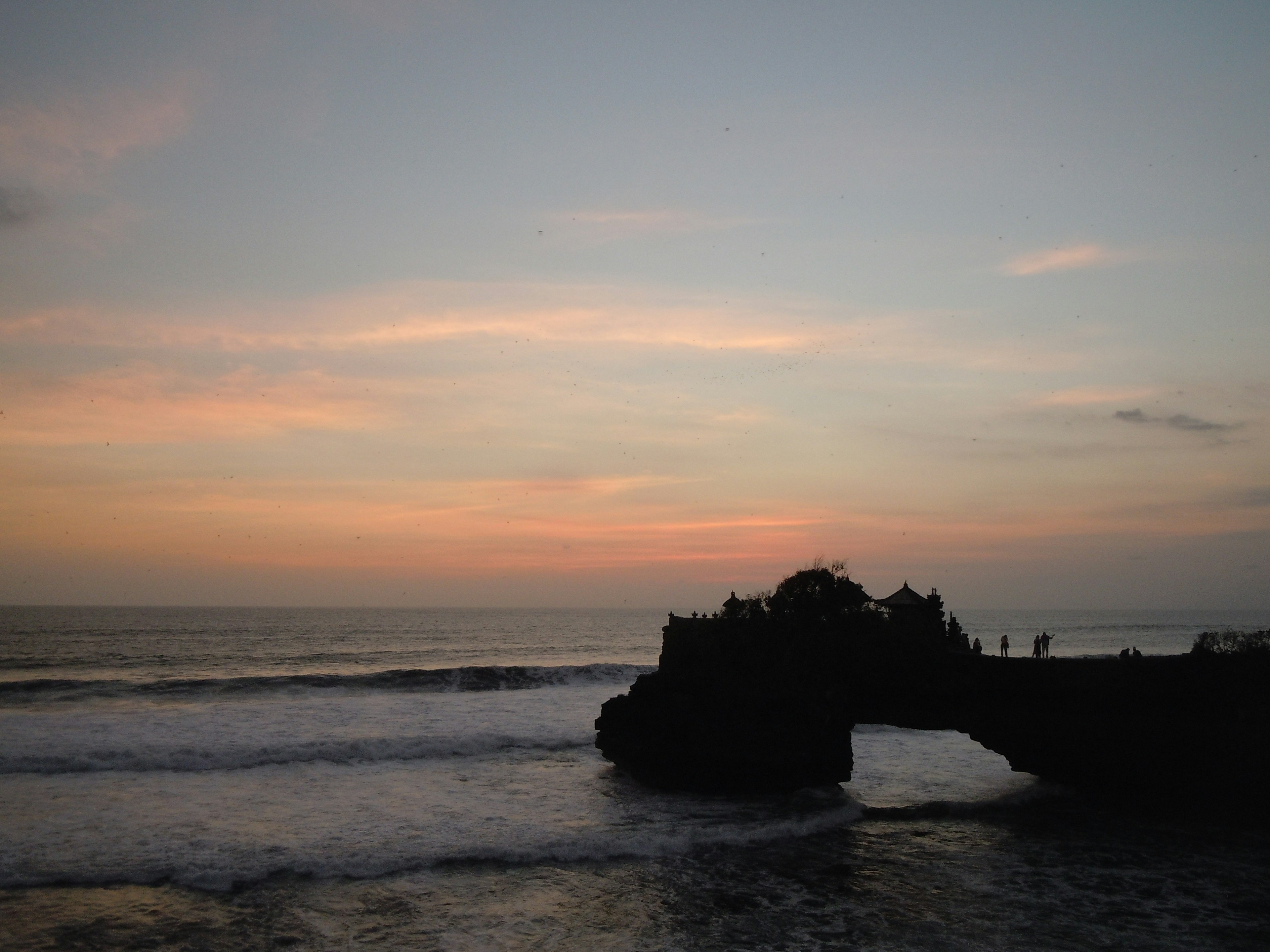 Pemandangan pantai saat senja dengan ombak menghantam batu dan awan oranye di langit