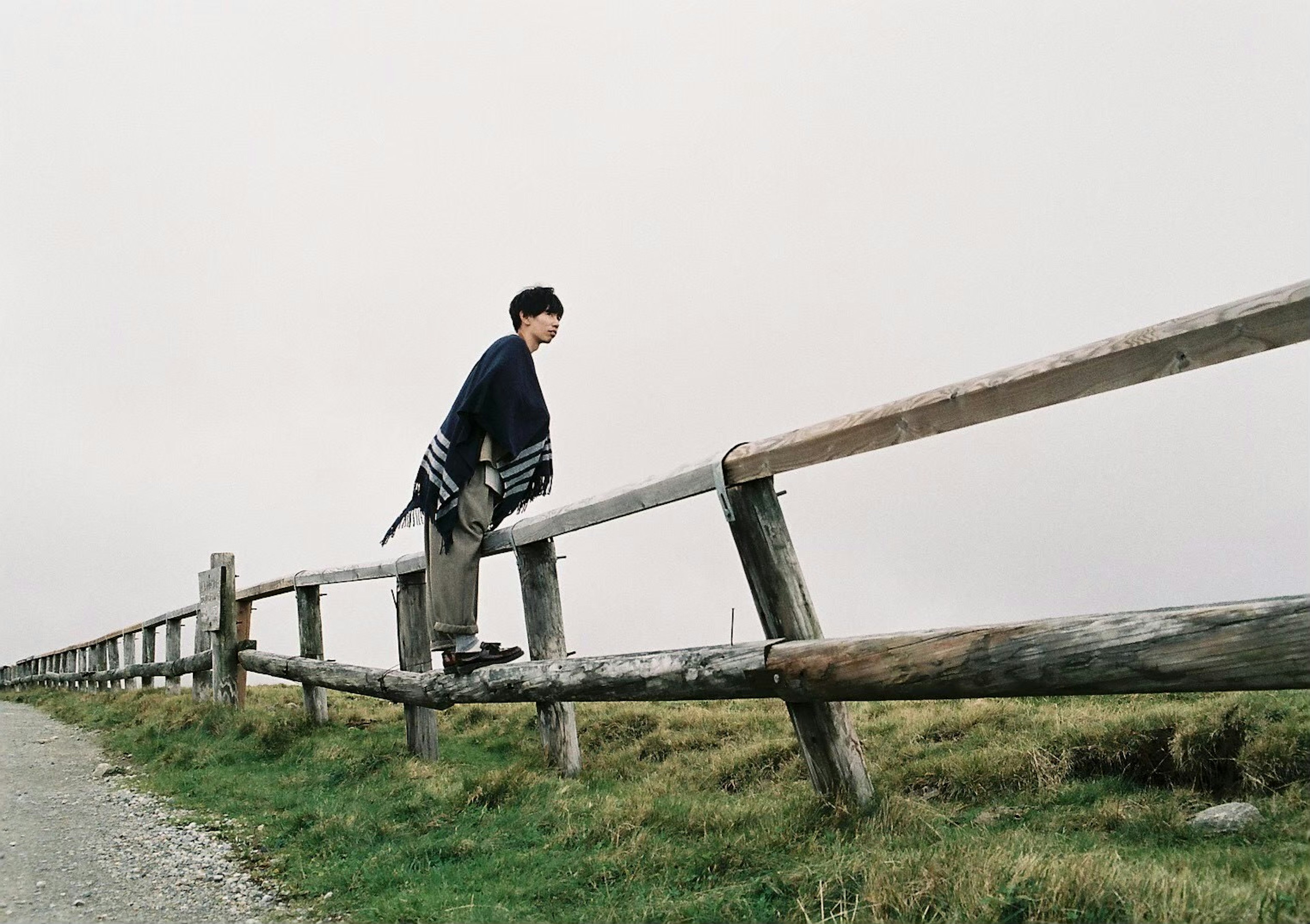 A man sitting on a wooden fence with a cloudy sky and green grassland in the background