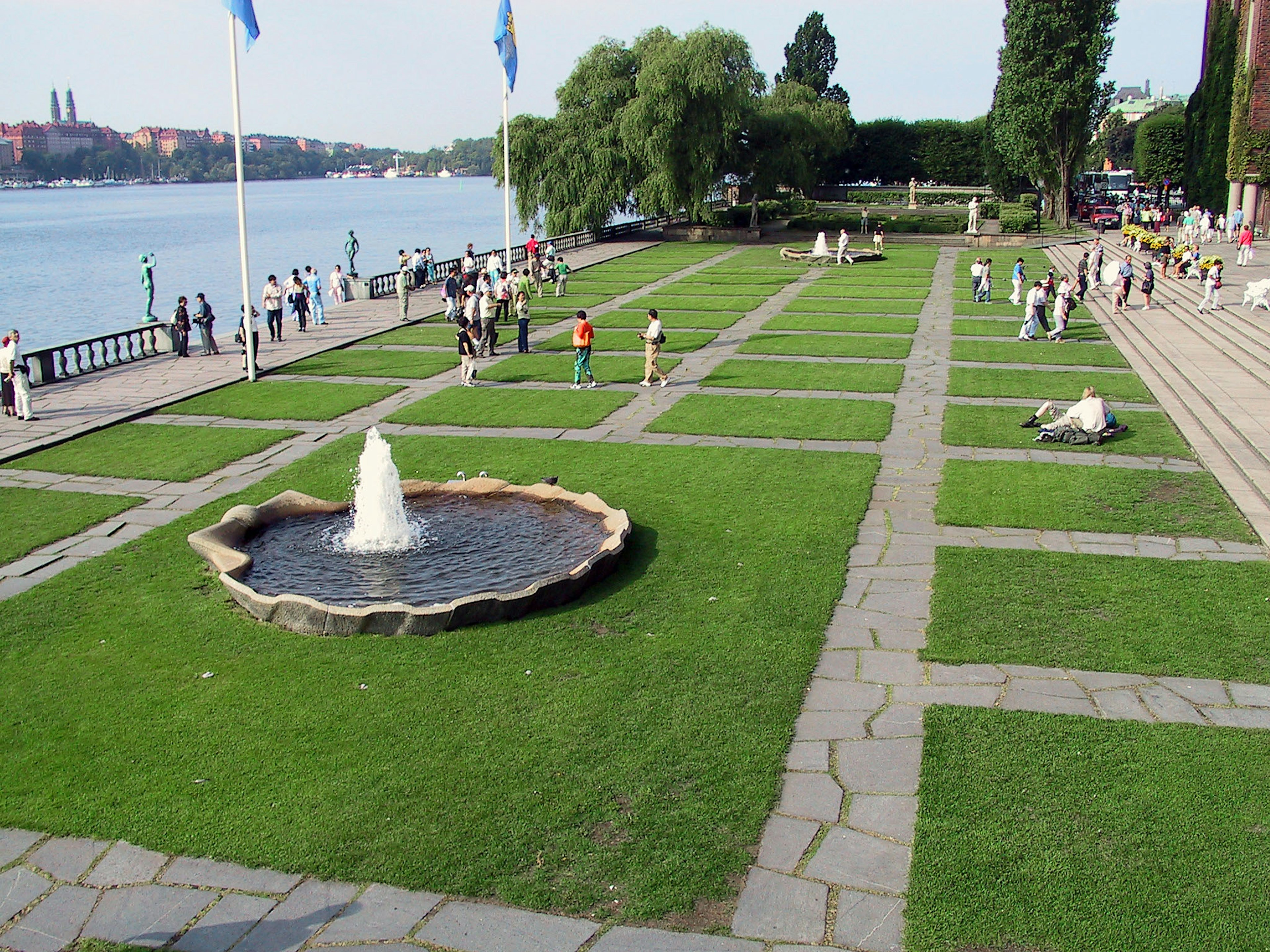 Parklandschaft mit Gras und Brunnen Menschen spazieren entlang eines Flusses