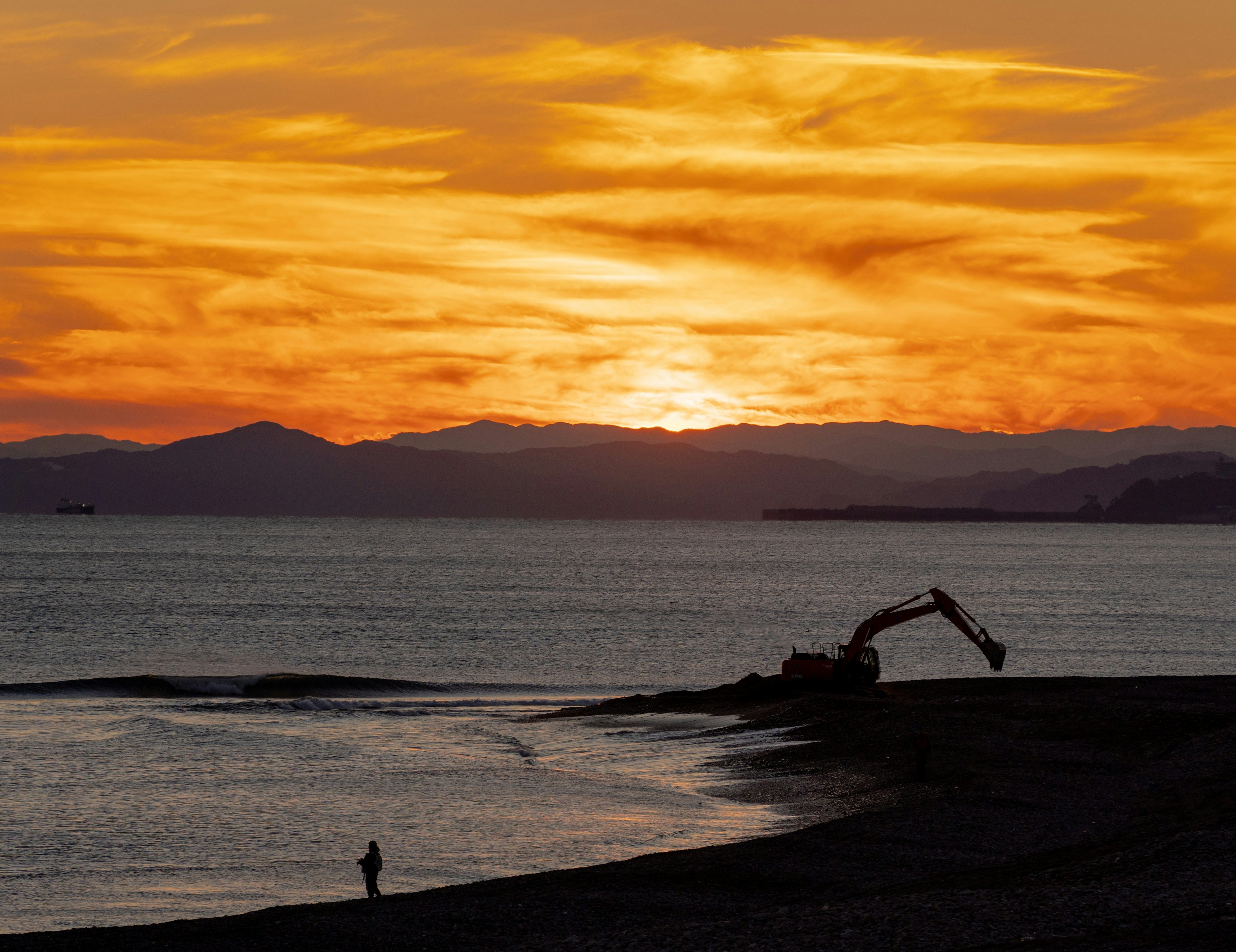 夕日が海を照らす美しい風景に、海岸で作業する重機と小さな人影が見える