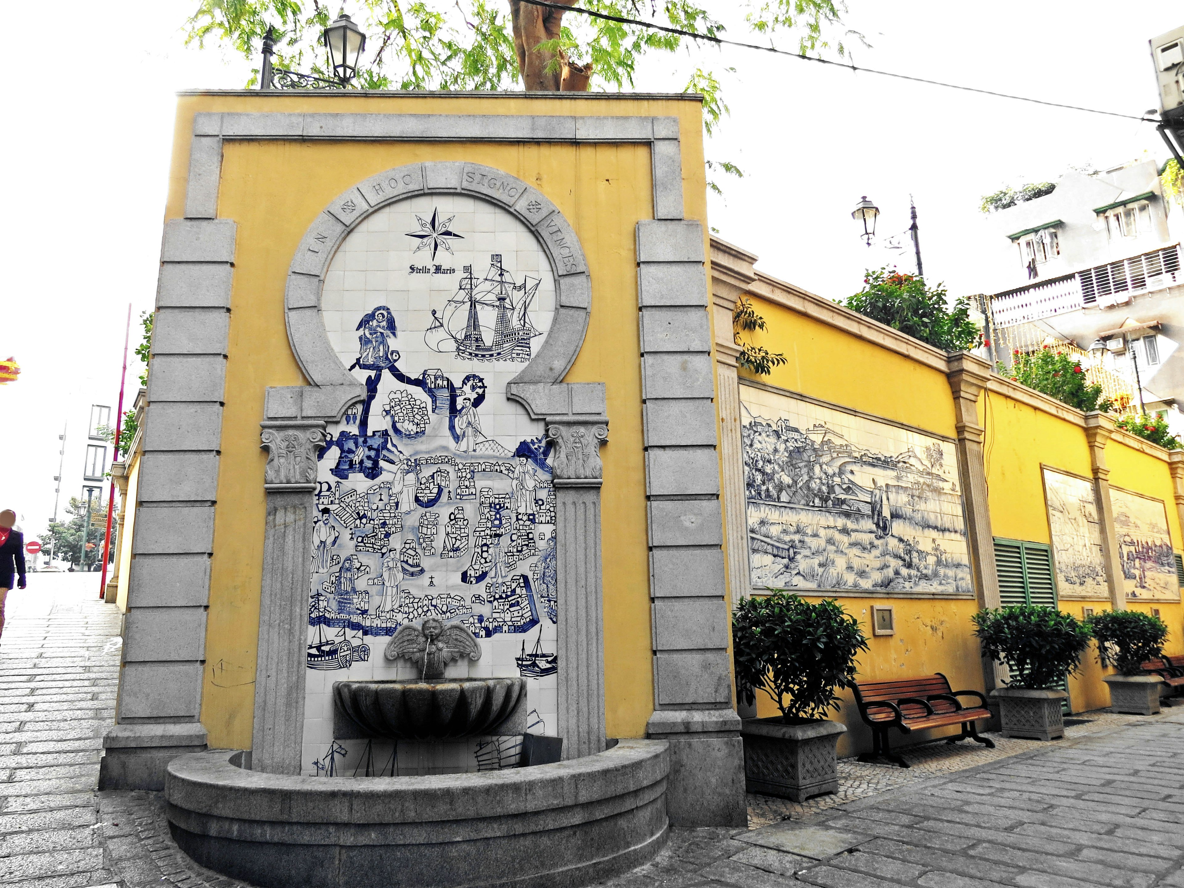 Street view featuring a fountain adorned with blue and white tiles against a yellow wall