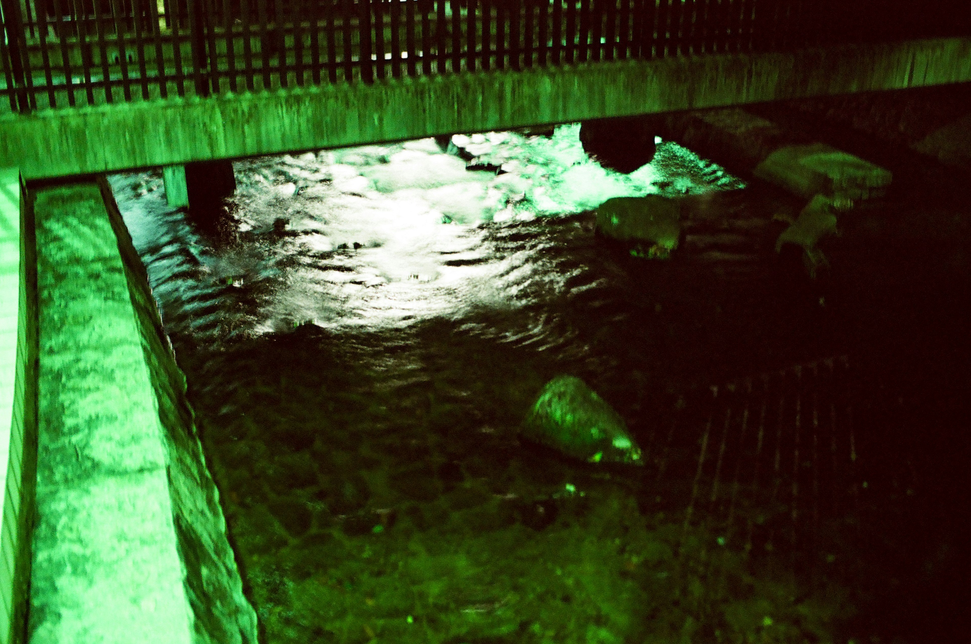 Under a bridge with flowing water illuminated by green light and visible rocks