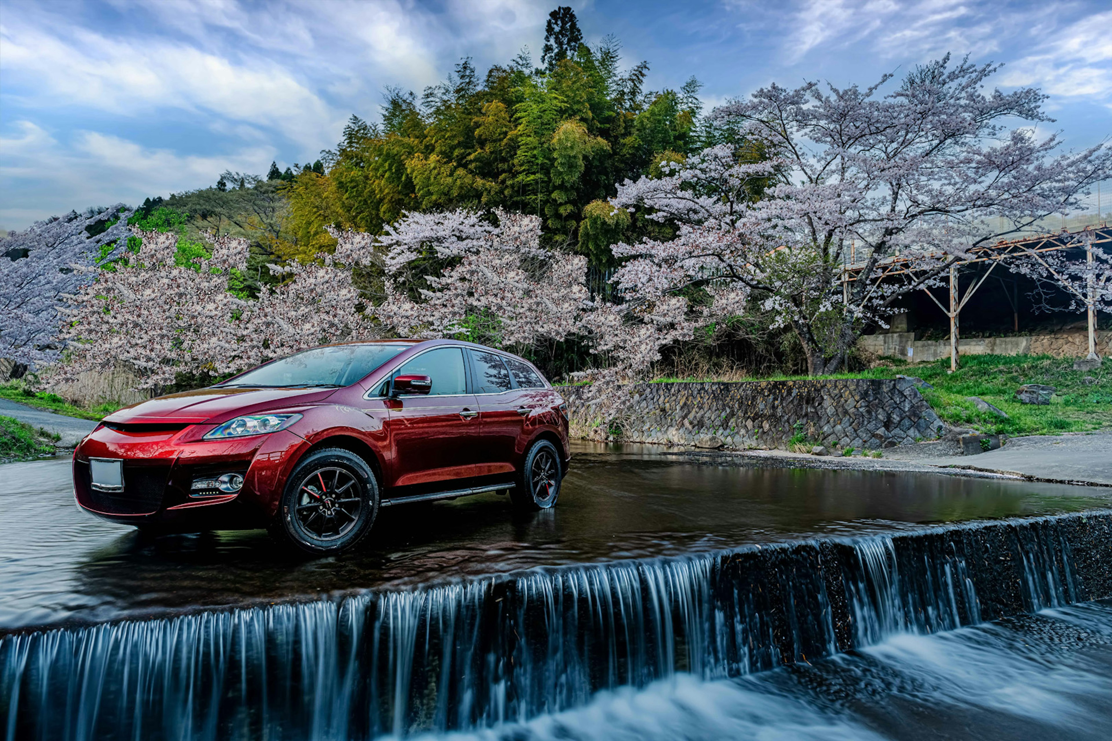 SUV merah diparkir di depan air terjun yang tenang dengan pohon sakura di latar belakang