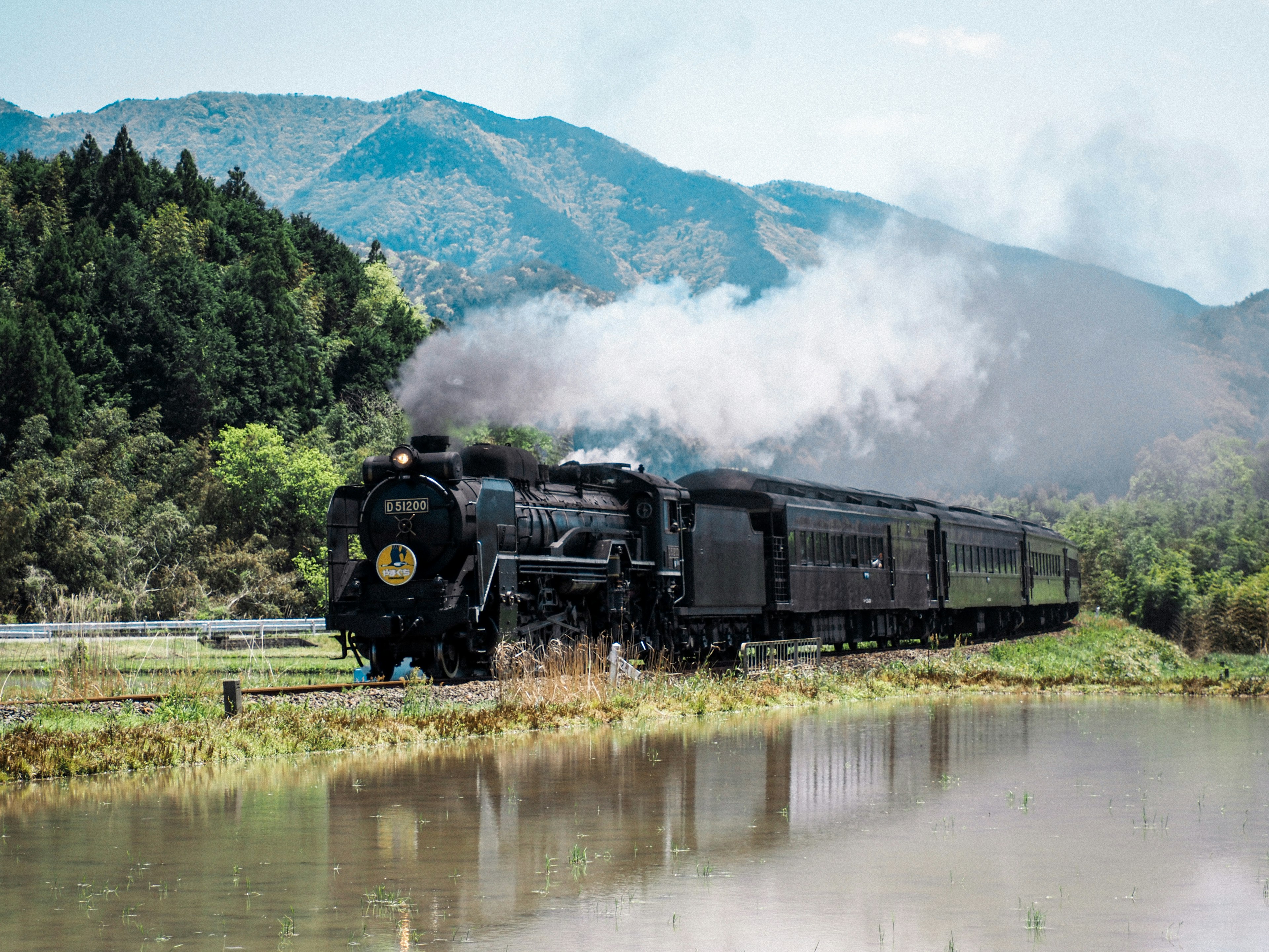 A steam locomotive traveling alongside a river with mountains in the background