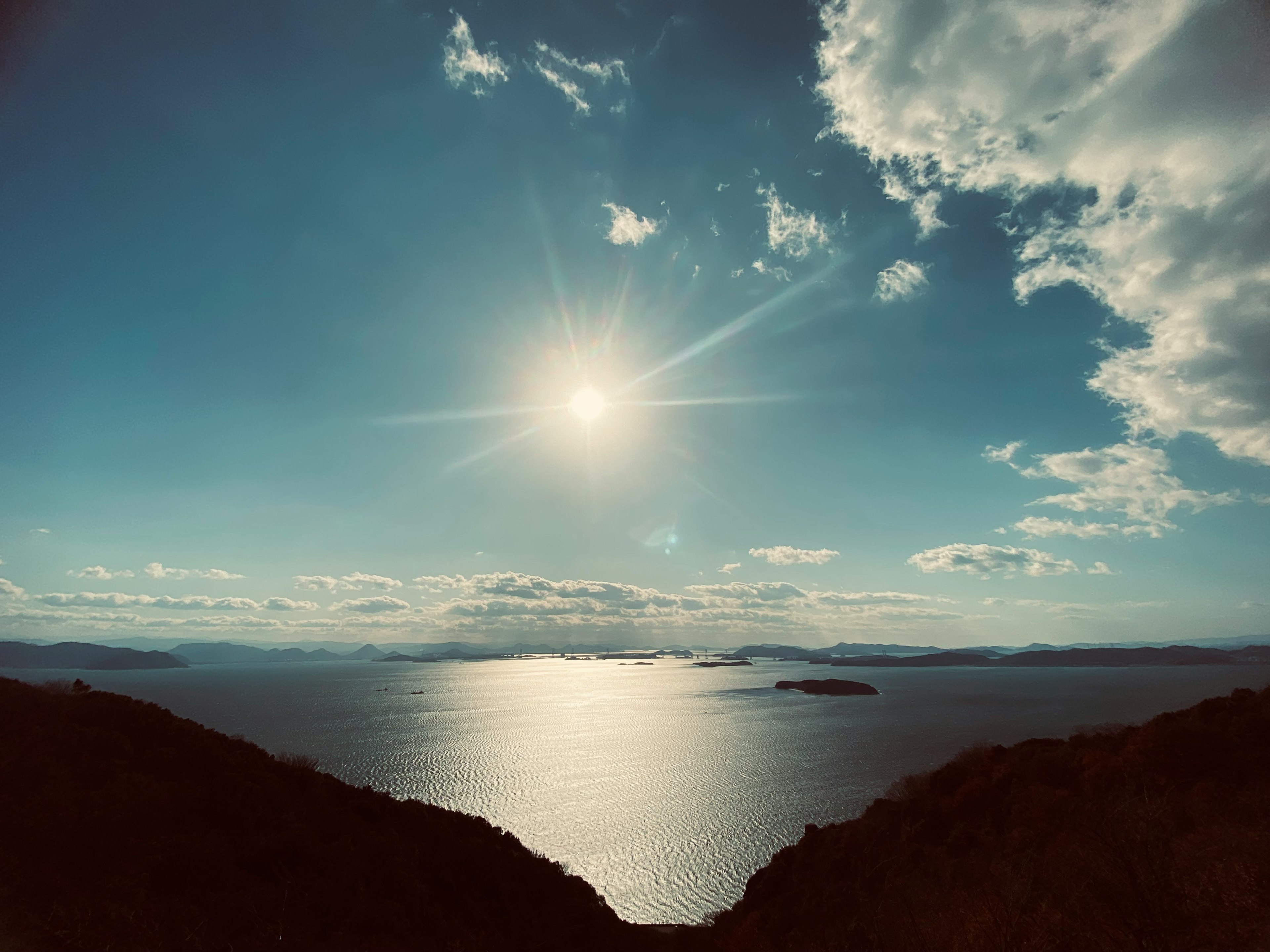 A scenic view of the sun shining over a calm sea under a blue sky with clouds