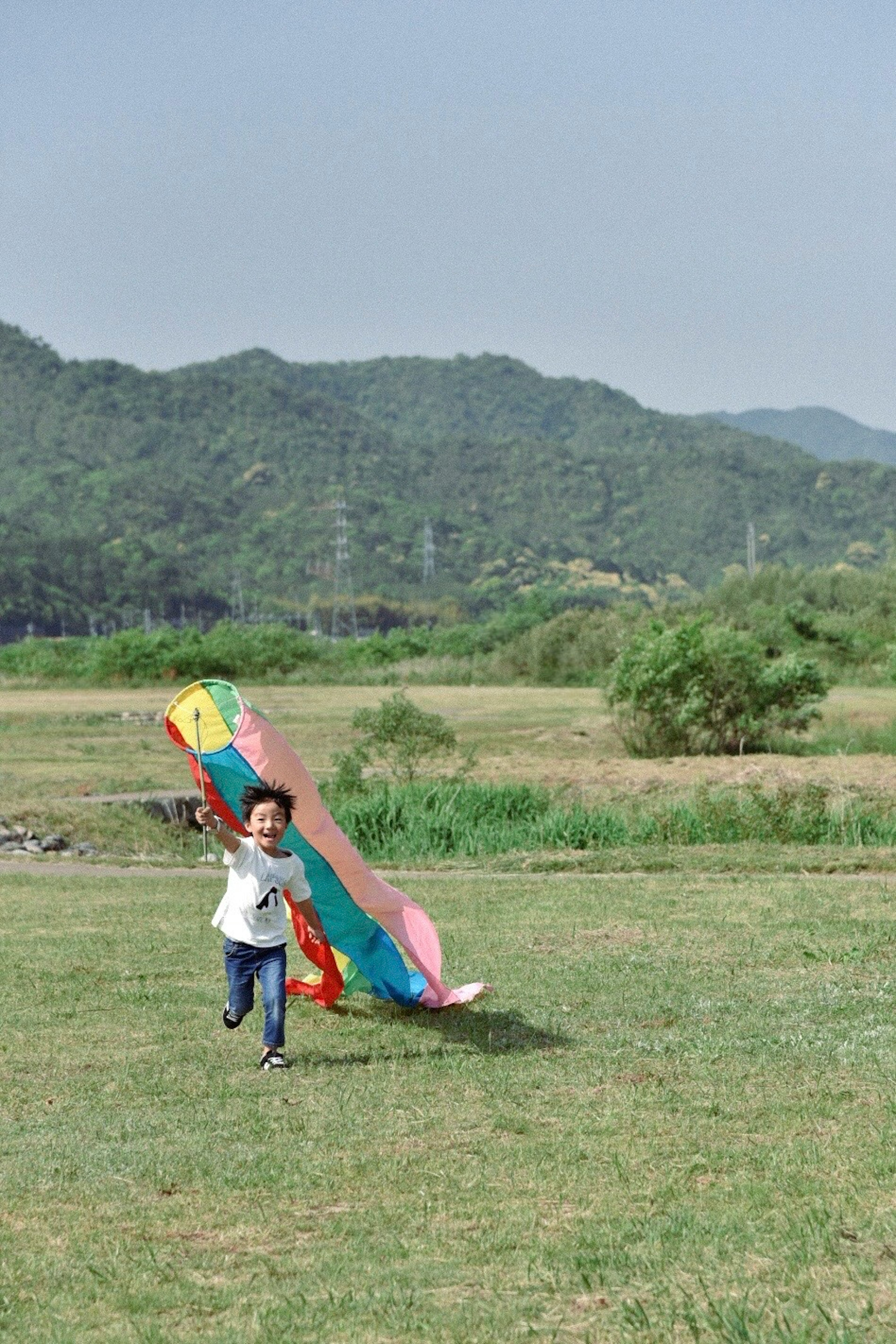 Child running with a colorful kite in a grassy field surrounded by mountains