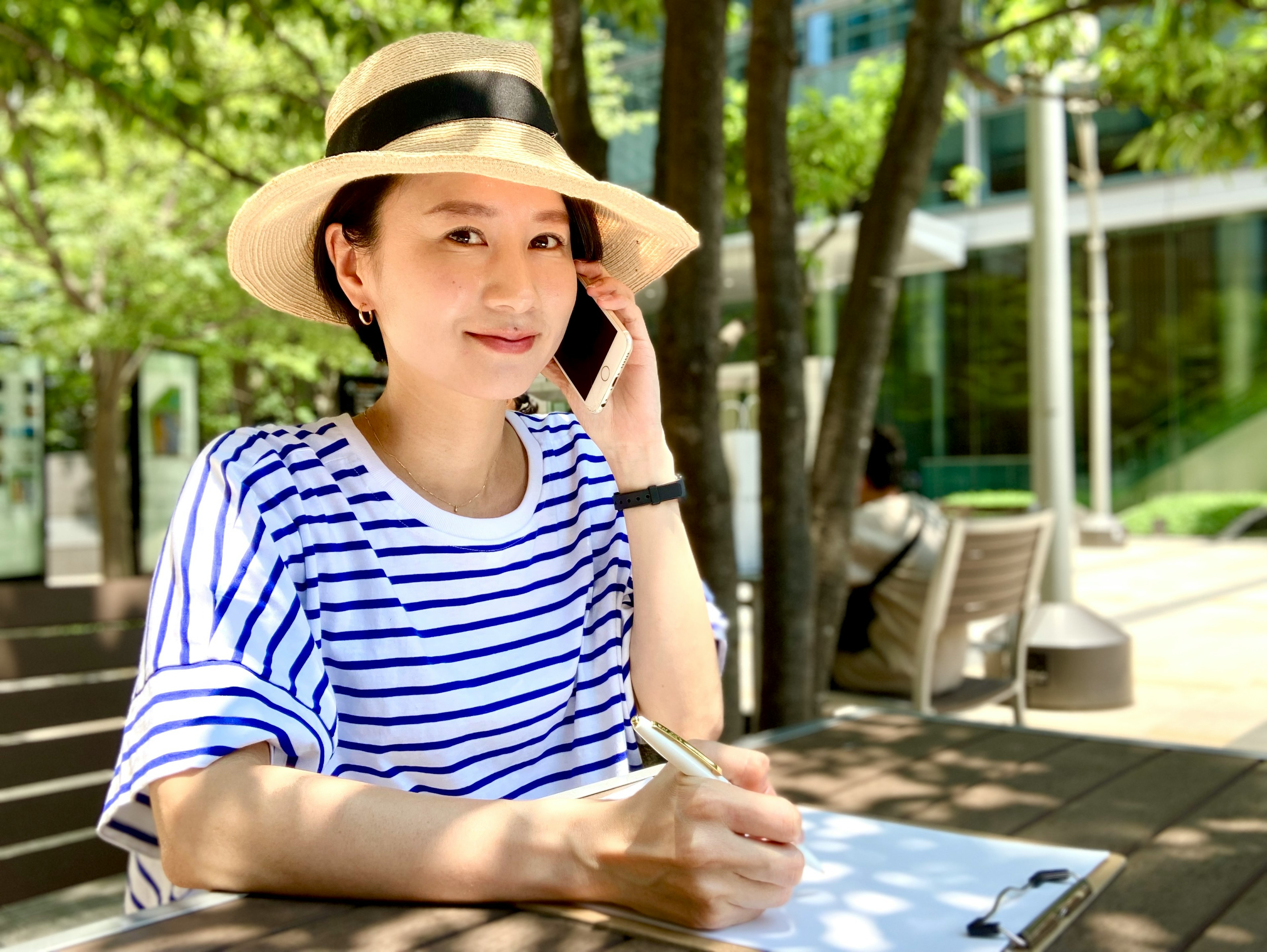 A woman in a striped shirt wearing a hat is talking on the phone while writing in a notebook