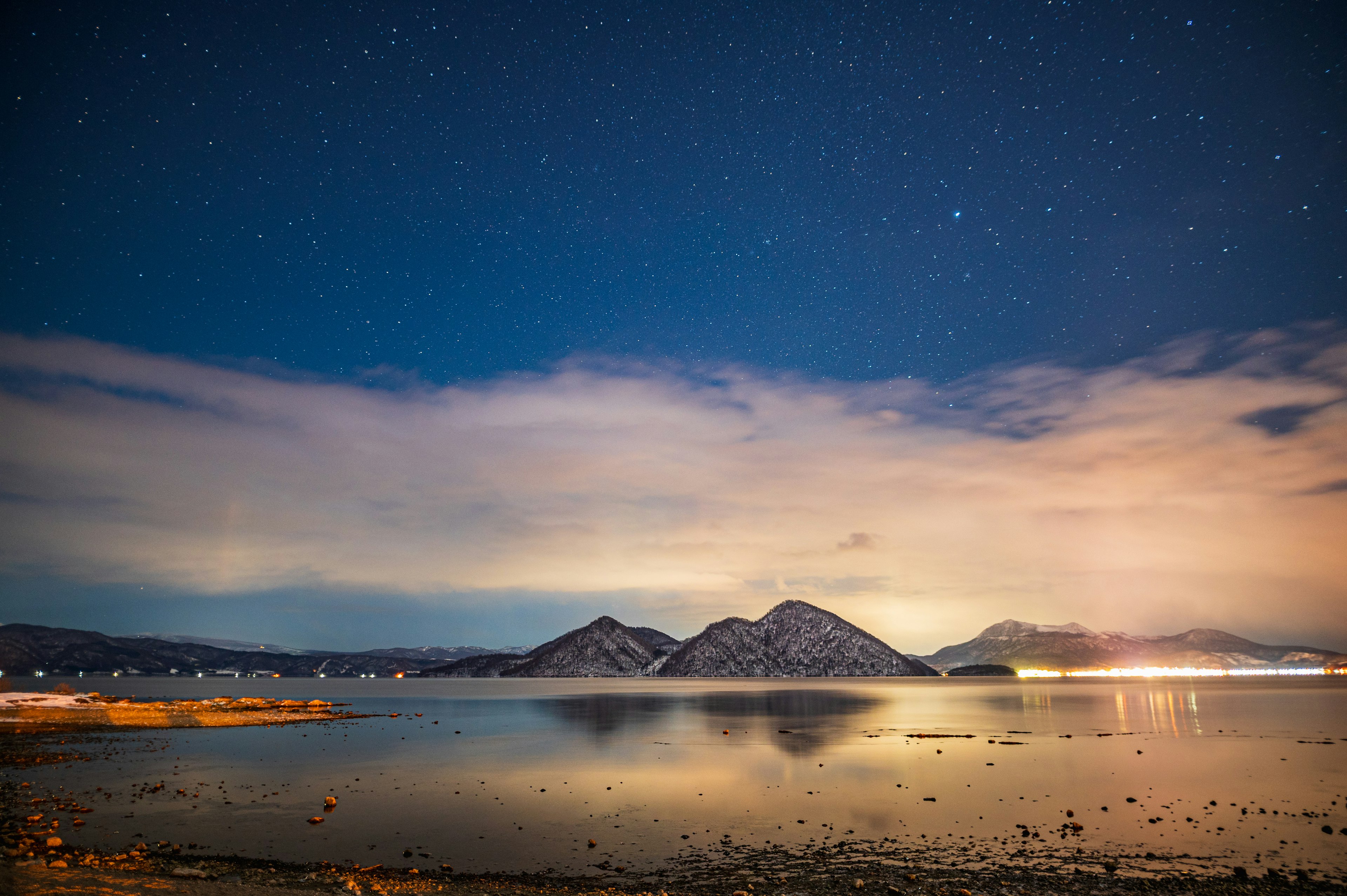 Langit malam berbintang di atas danau tenang dengan pegunungan berkabut di latar belakang